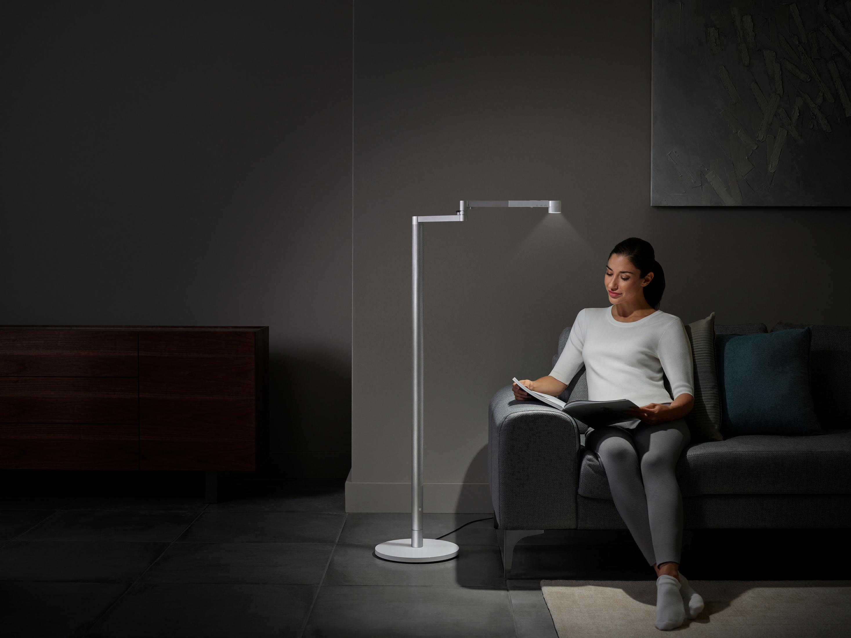 Eine Frau liest auf einem Sofa unter einer Stehlampe. Dunkler Raum, graues Sofa und Licht von der Lampe.