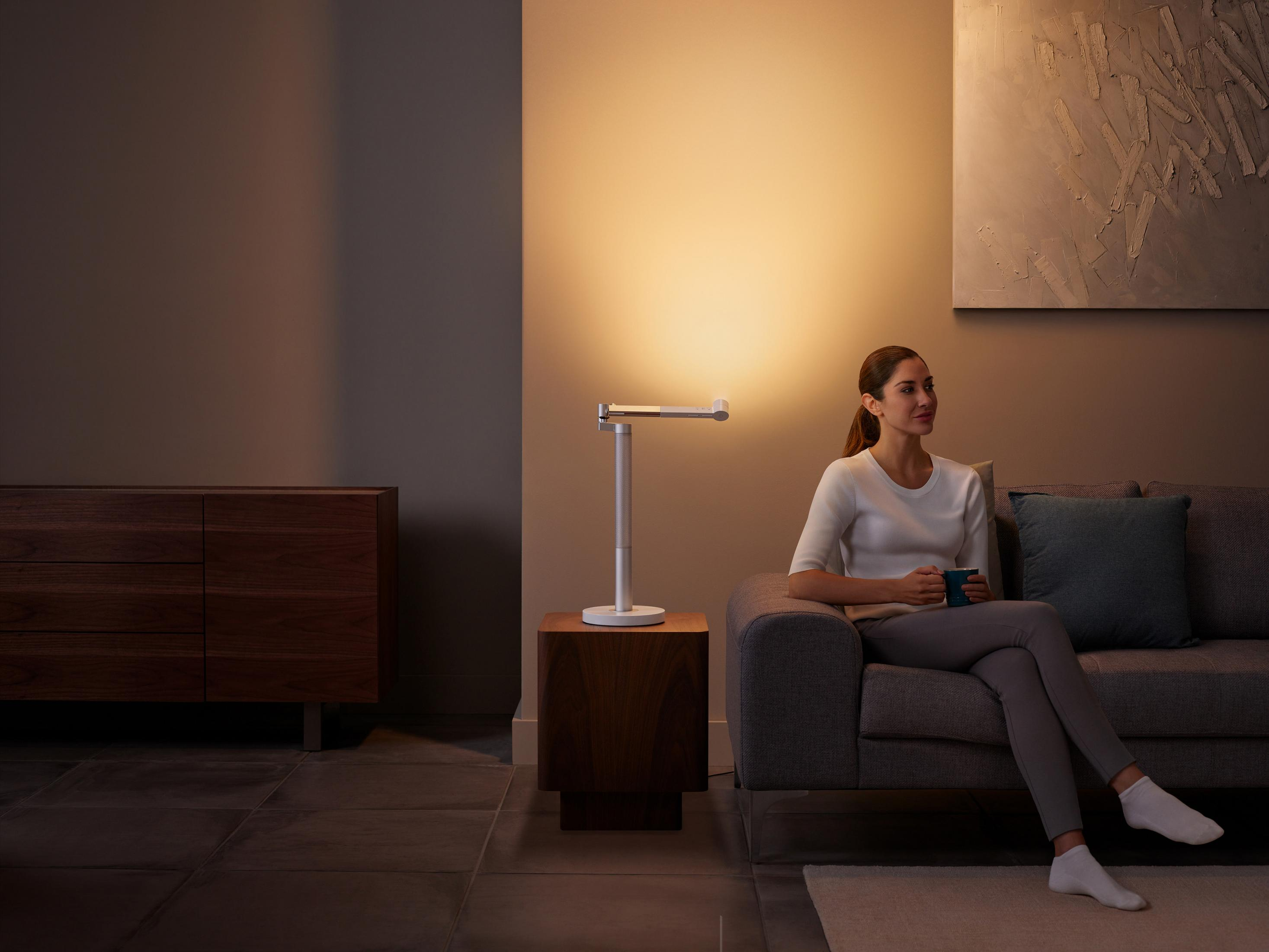 Frau sitzt auf einem Sofa, von einer Lampe beleuchtet, hält einen Becher, mit einem Beistelltisch.