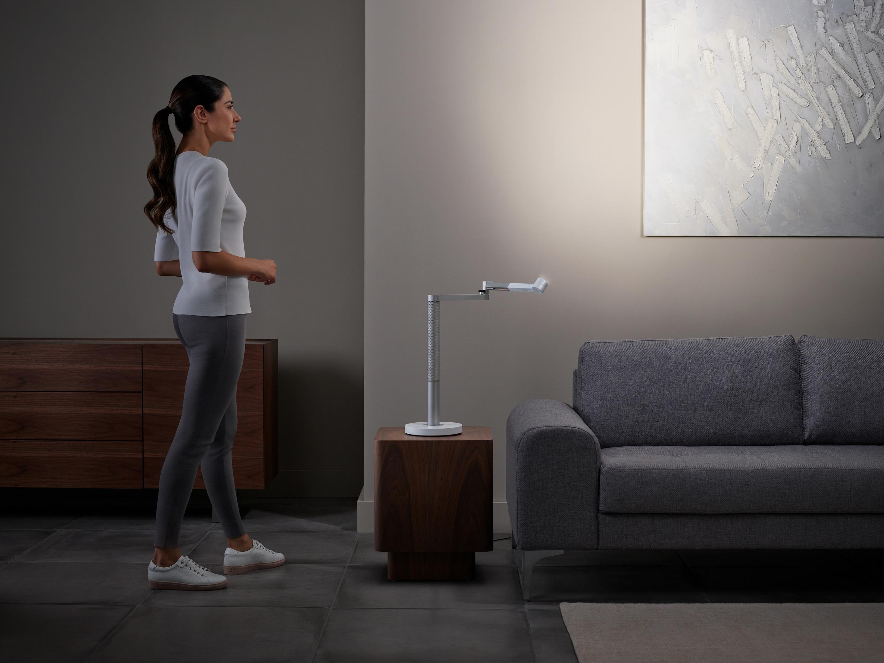 Frau geht in der Nähe einer modernen Lampe auf einem Holzständer, graues Sofa und Gemälde an der Wand.