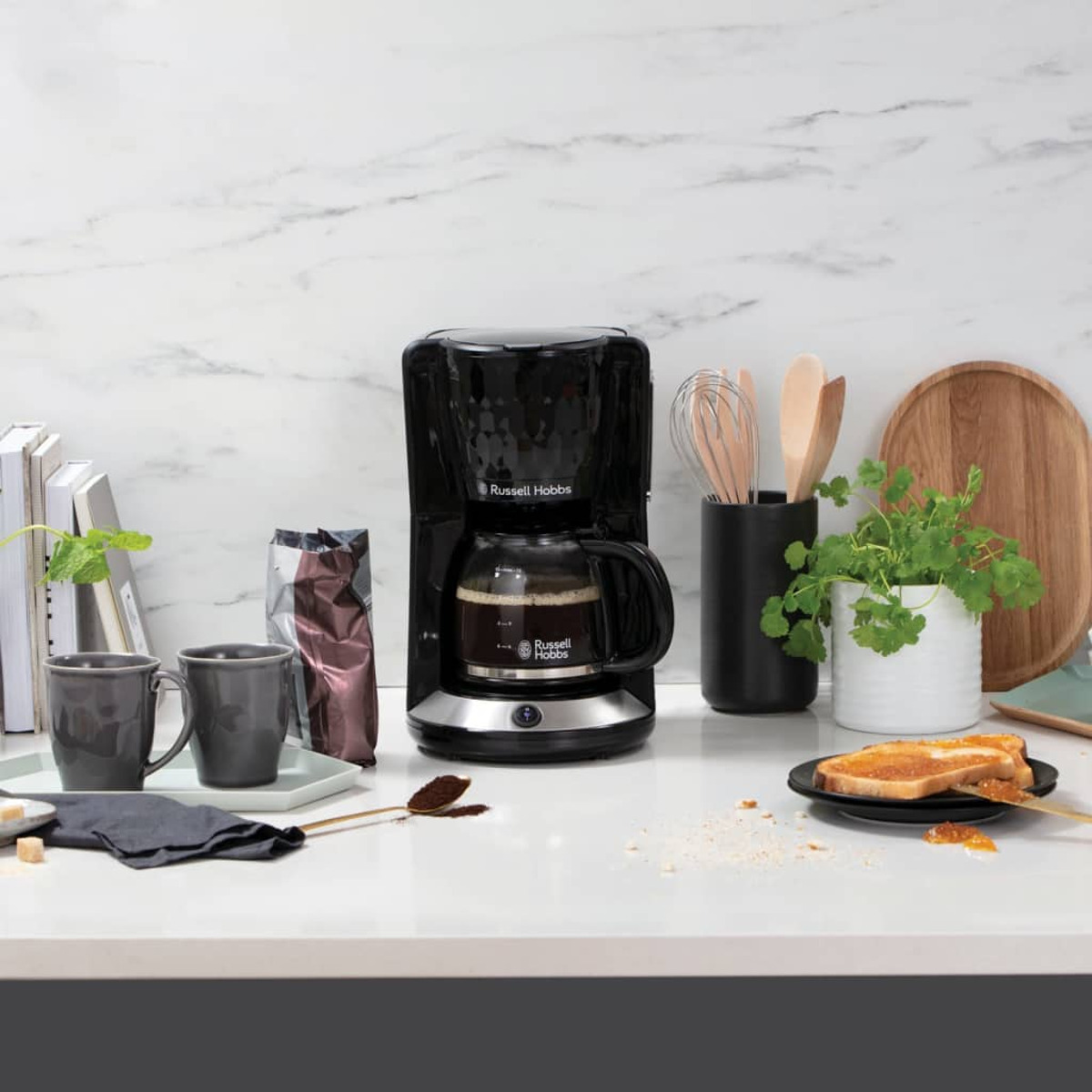 Una cafetera negra, tazas de café, una planta y tostadas en una encimera blanca.