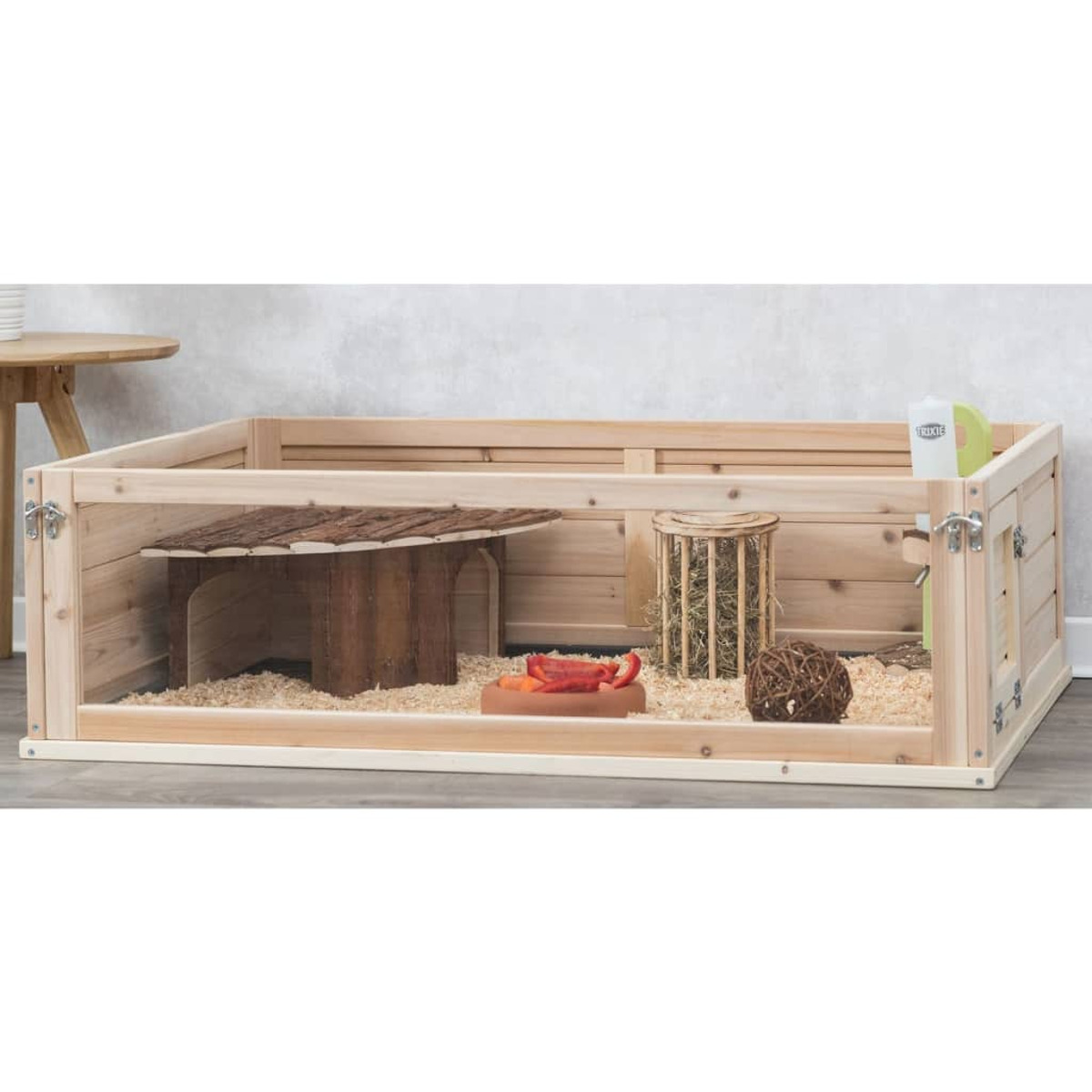 Corral para mascotas de madera que contiene virutas de madera, comida y una botella de agua.