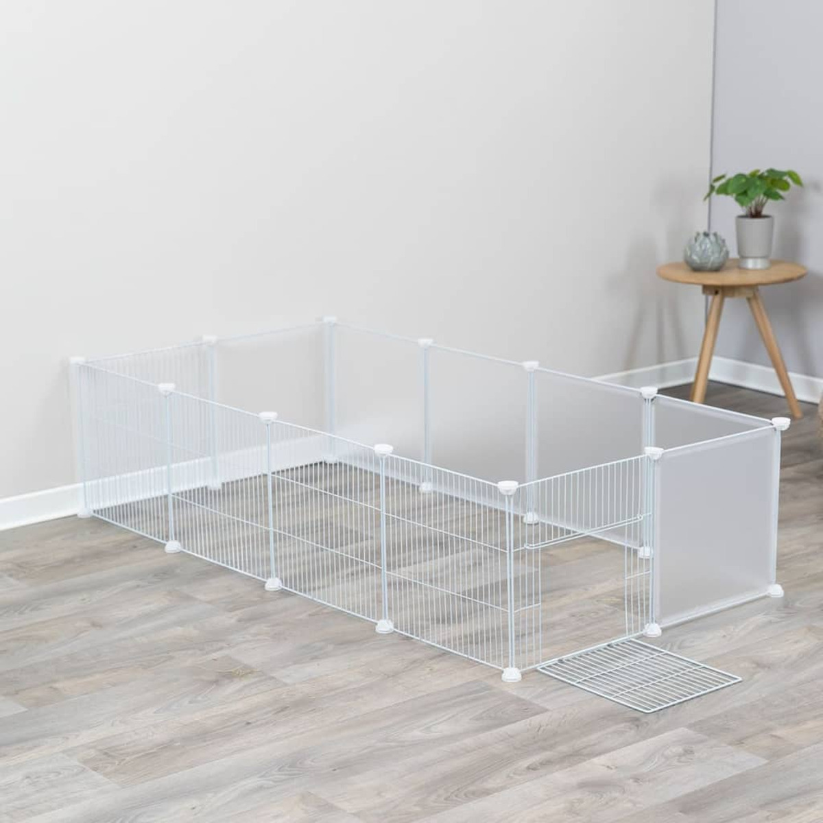 Weißes Haustiergehege mit Drahtgeflecht und Kunststoffplatten. Grauer Boden, neutrale Wand und kleiner Tisch hinten.