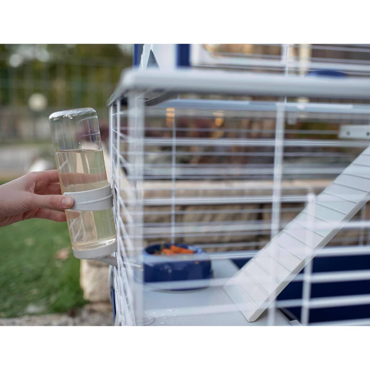 Mano sosteniendo una botella de agua junto a una jaula para animales. Rampa en el fondo.