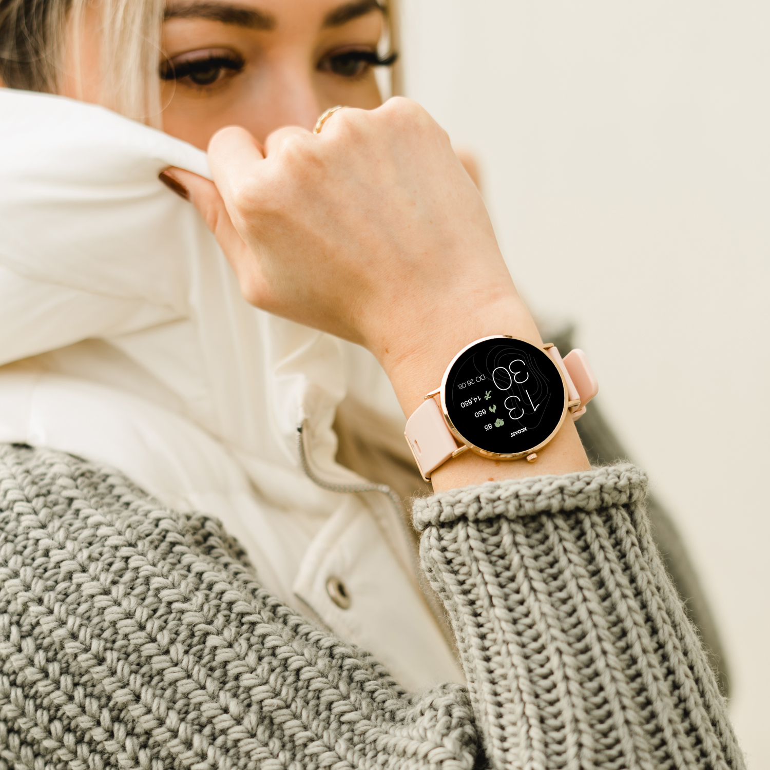 Frau mit grauem Pullover und Smartwatch mit rosa Armband, die die Zeit anzeigt.