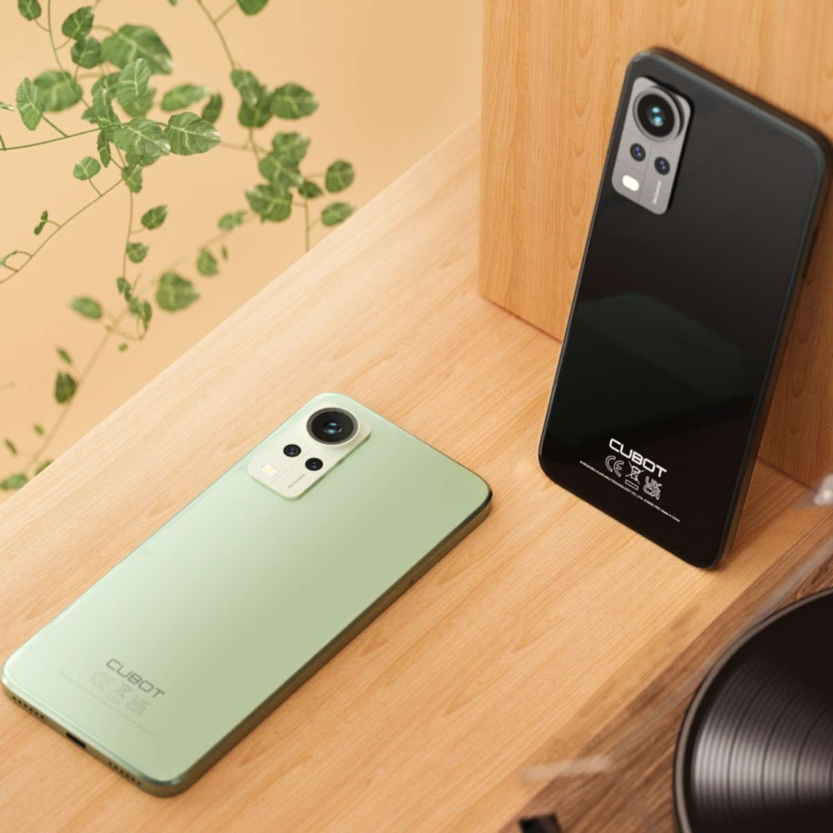 Dos smartphones, verde y negro, sobre una superficie de madera con una planta al fondo.