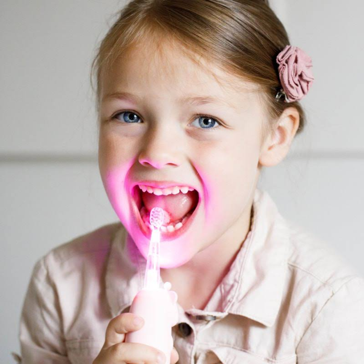 Una bambina con la bocca aperta, che tiene uno spazzolino rosa luminoso. Luce rosa.