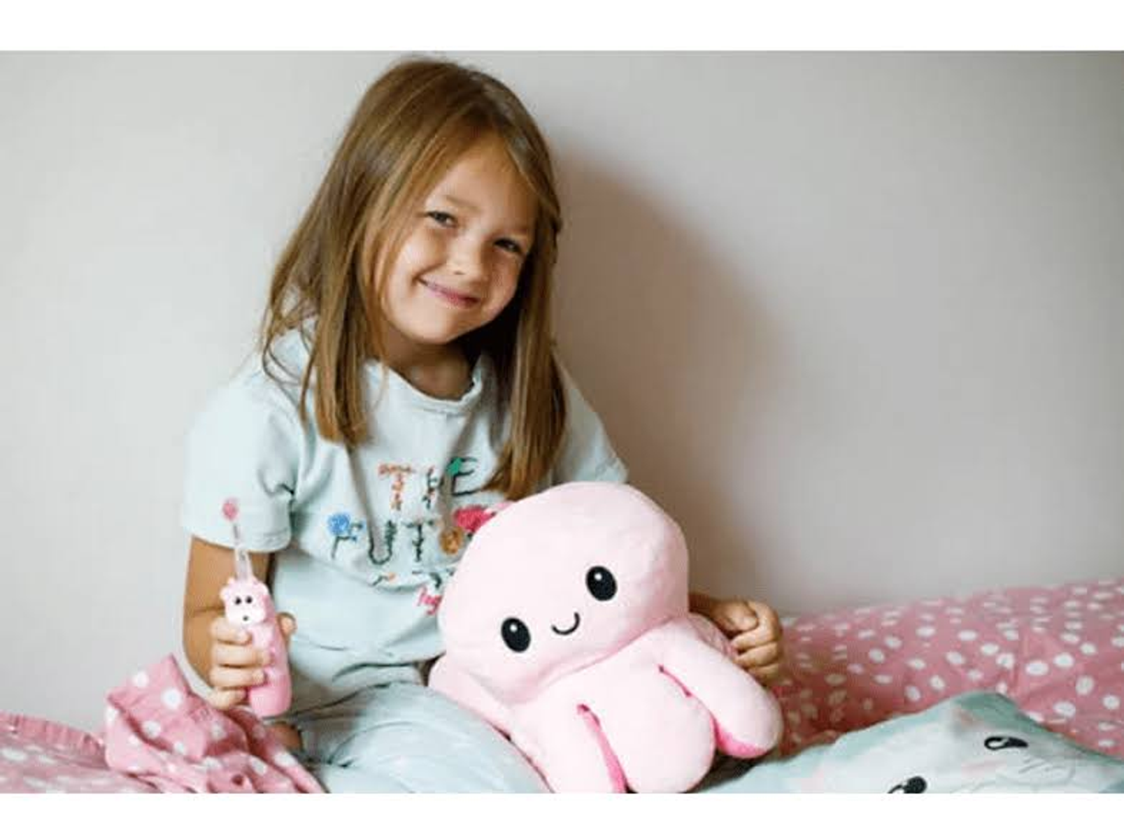 Una bambina sorridente, con un peluche di polpo rosa e uno spazzolino da denti rosa.