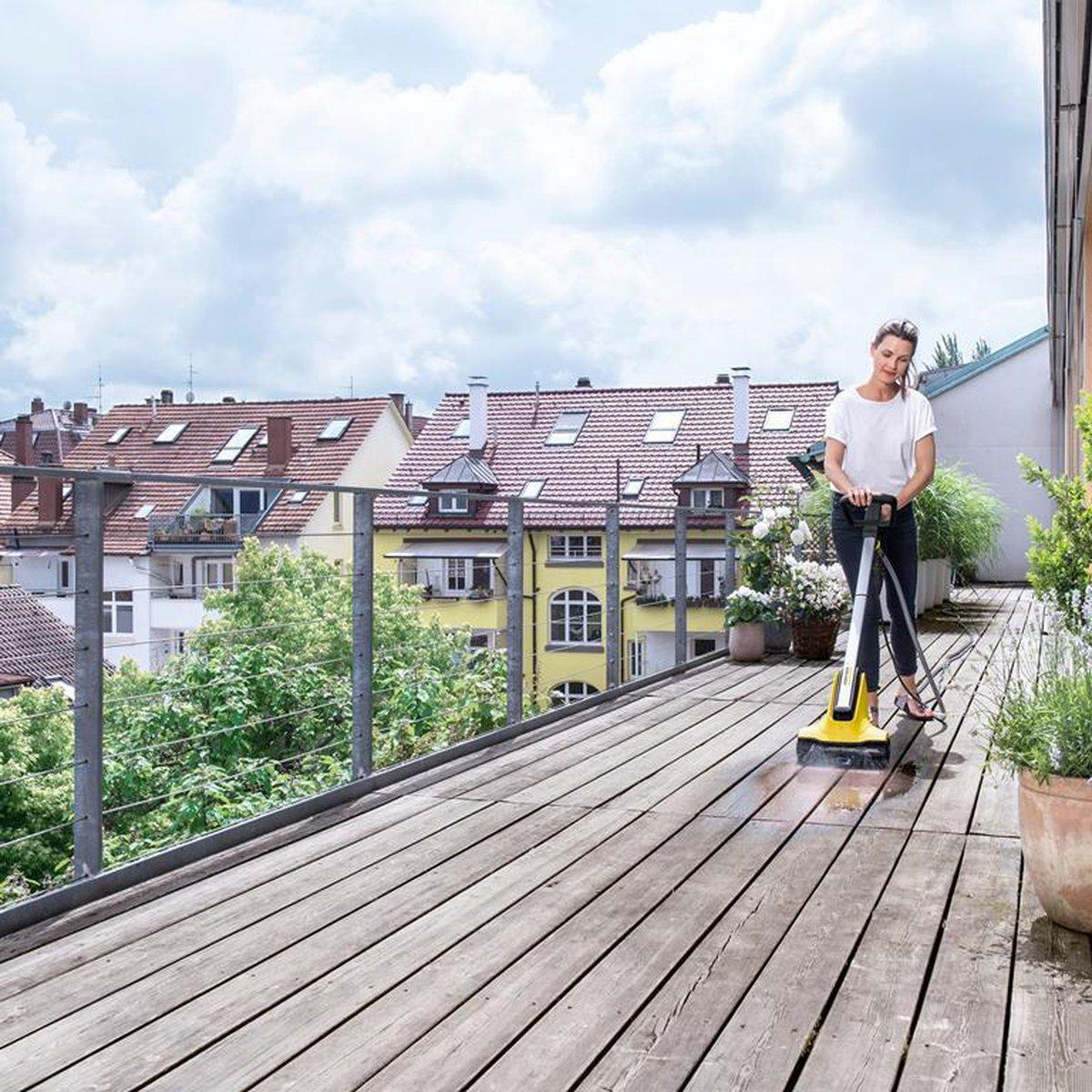 Kobieta czyści drewniany taras żółtym urządzeniem. Budynki w tle.