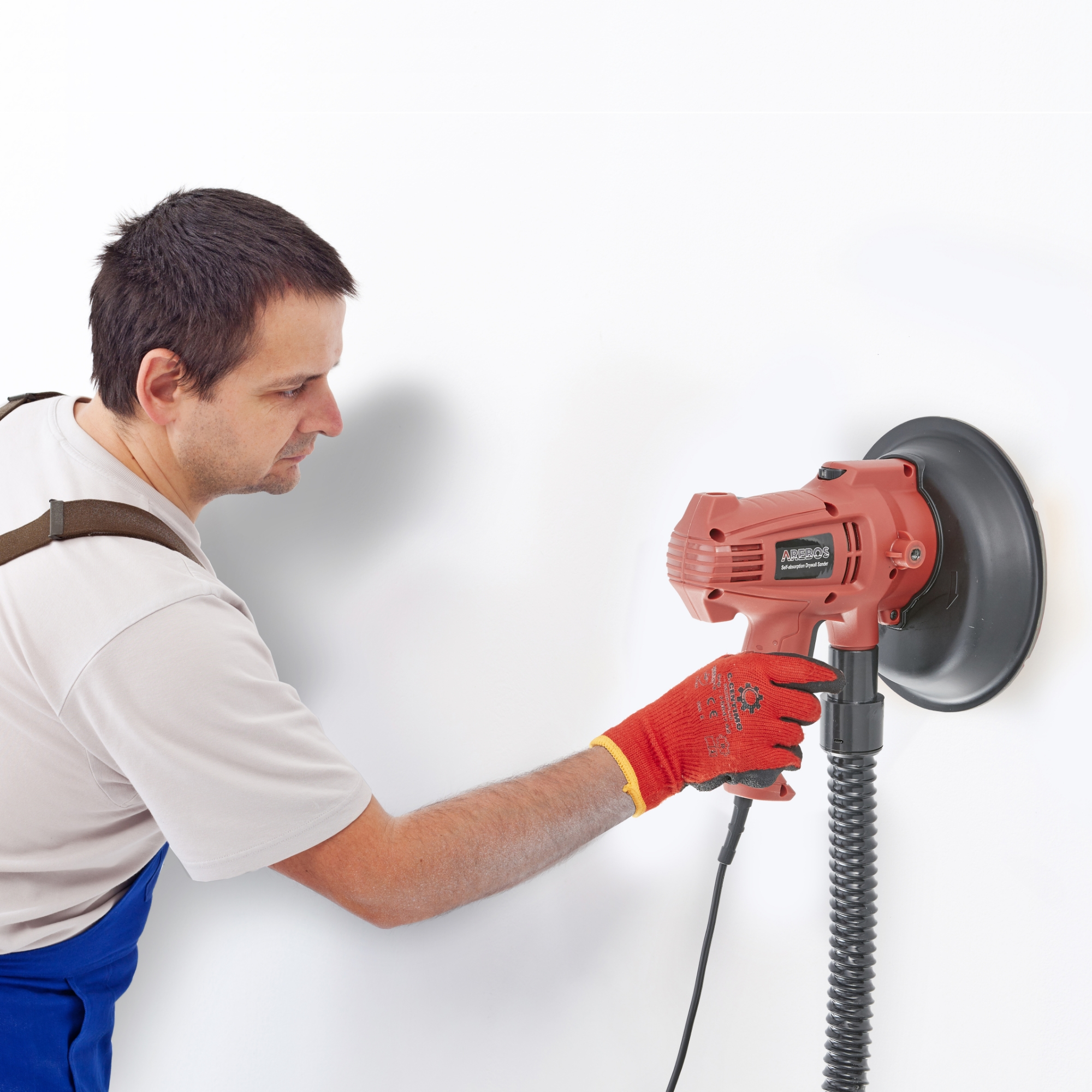 Ein Mann in blauen Overalls und roten Handschuhen benutzt einen roten Wandschleifer gegen eine weiße Wand.