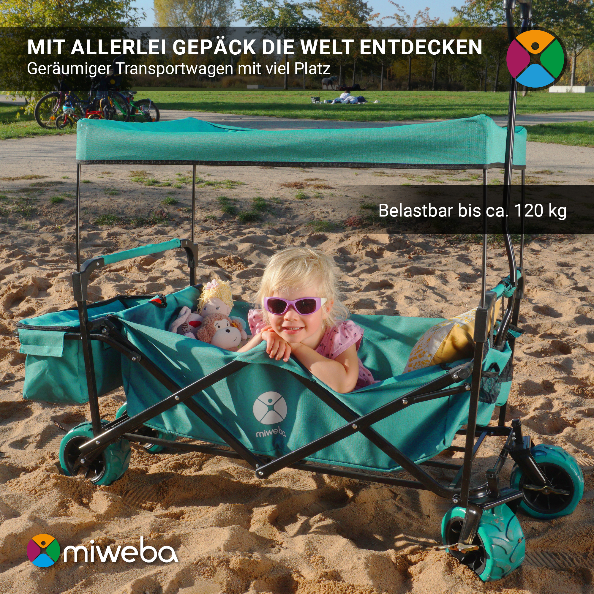 Ein Kind in einem blauen Wagen mit Spielzeug. Der Wagen hat ein grünes Verdeck an einem Sandstrand.