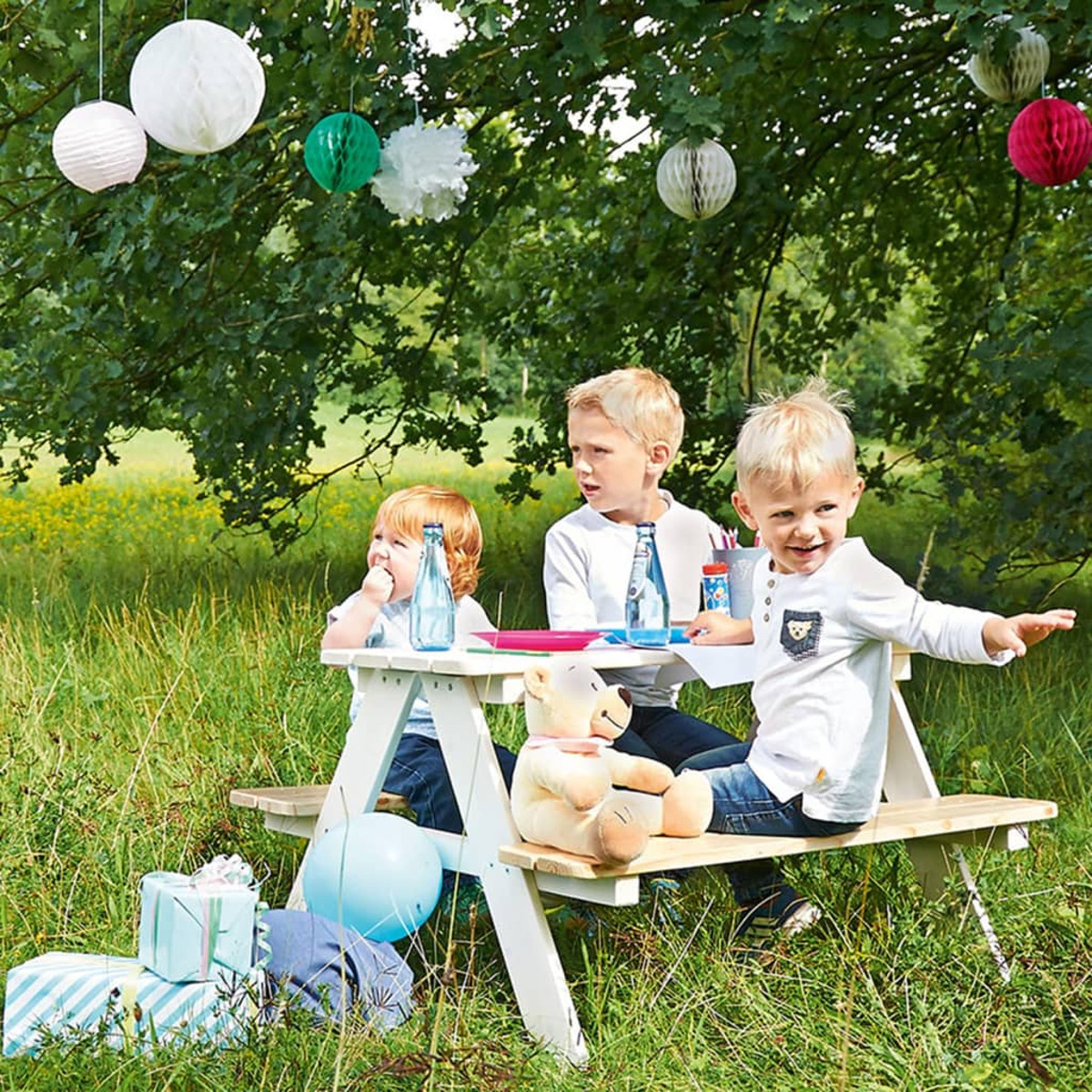Drei Kinder an einem Picknicktisch unter einem Baum mit Dekorationen.