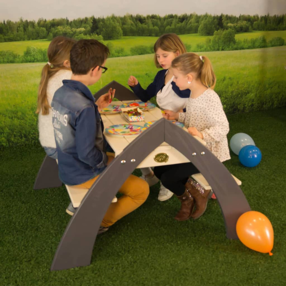 Vier Kinder an einem Picknicktisch. Grünes Gras und Luftballons im Hintergrund.