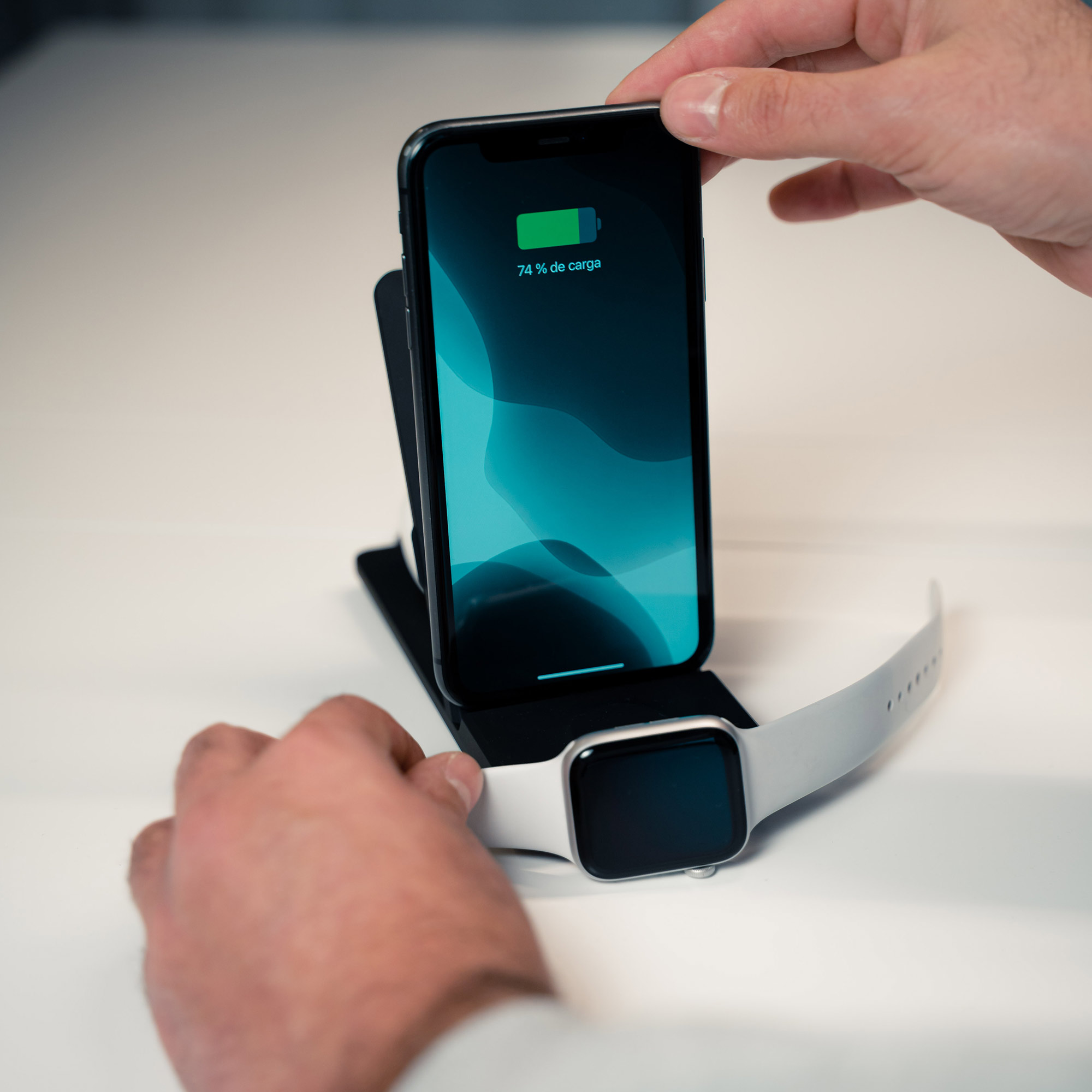 Manos sosteniendo un teléfono cargándose y un Apple Watch en un soporte de carga. Superficie blanca.