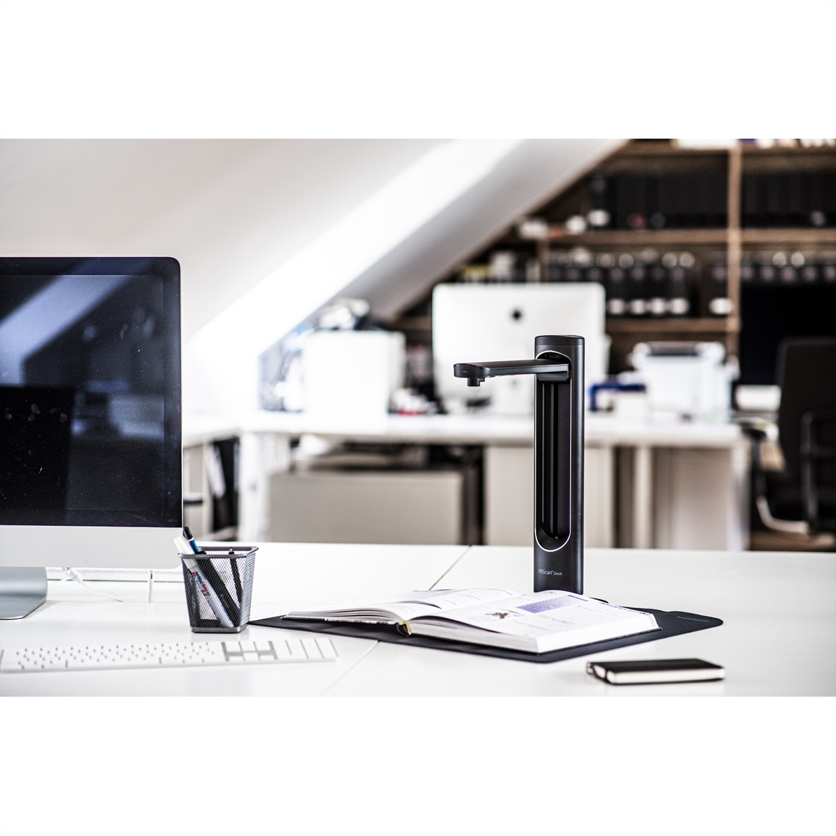 Un escáner de documentos en un escritorio blanco, con un monitor, teclado, bolígrafos y un cuaderno.