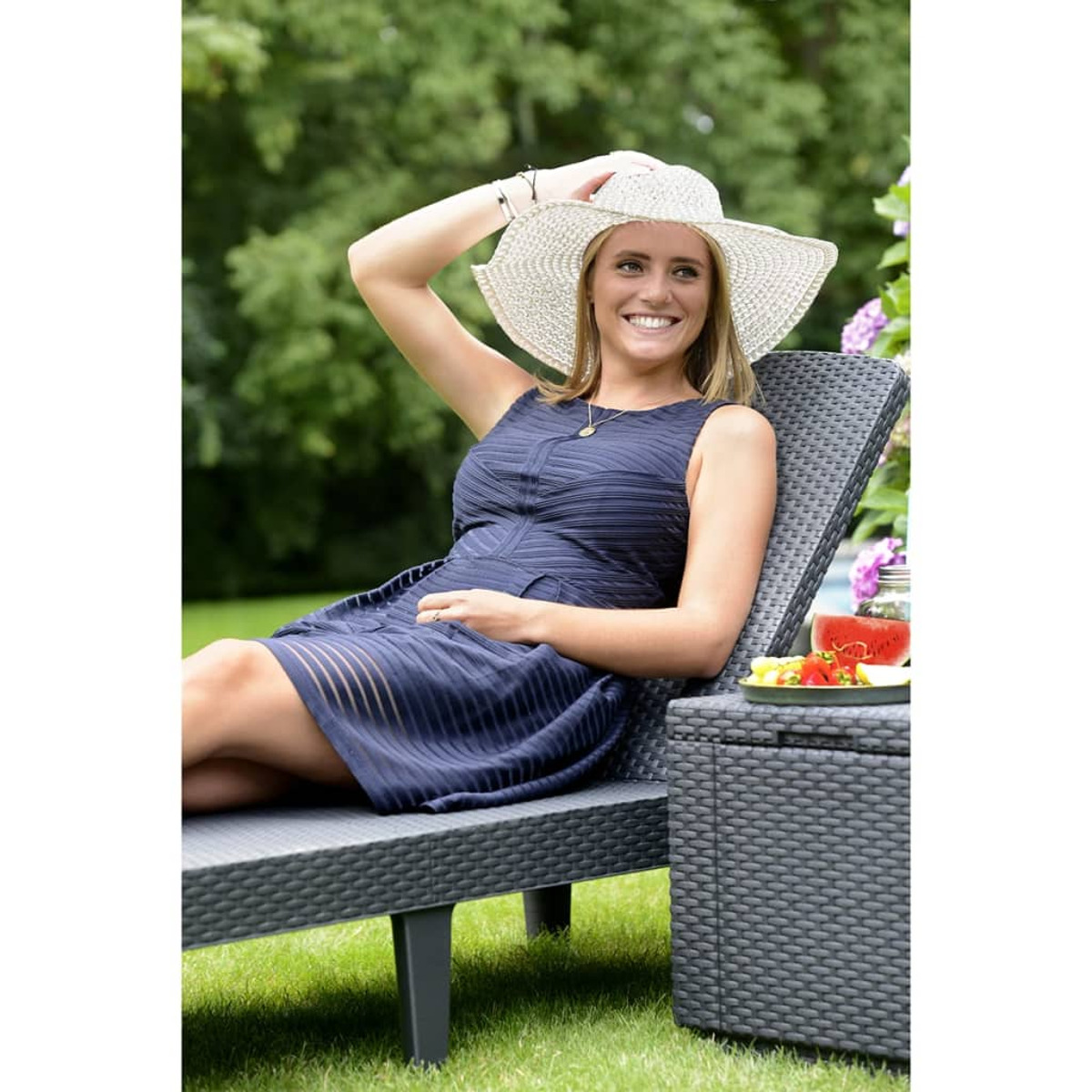 Donna in abito blu navy su una sedia a sdraio, sorridente. Accanto, un tavolino con cibo e bevande.