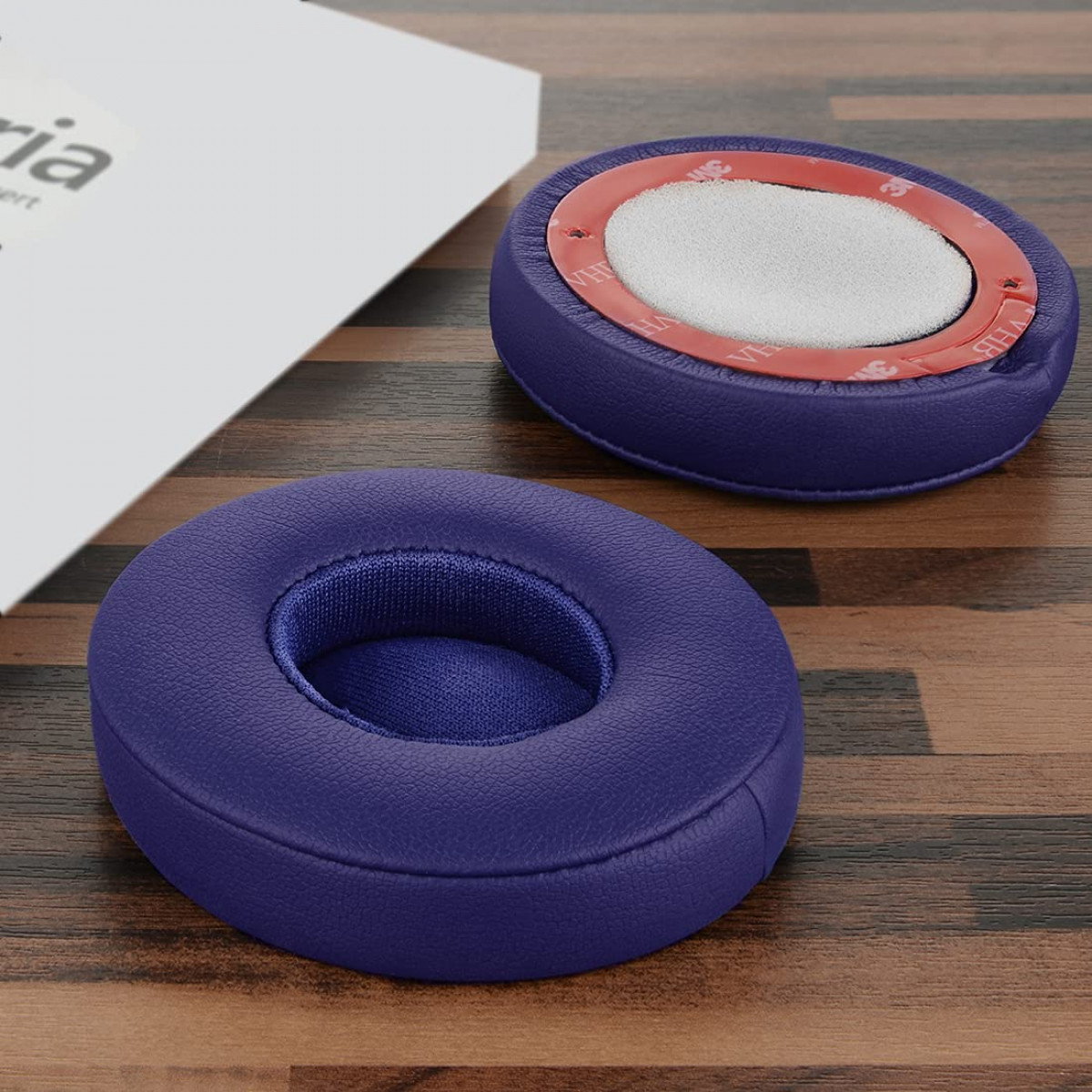 Zwei violette Kopfhörerpolster mit roten Kleberingen auf einer Holzoberfläche neben einer weißen Box.