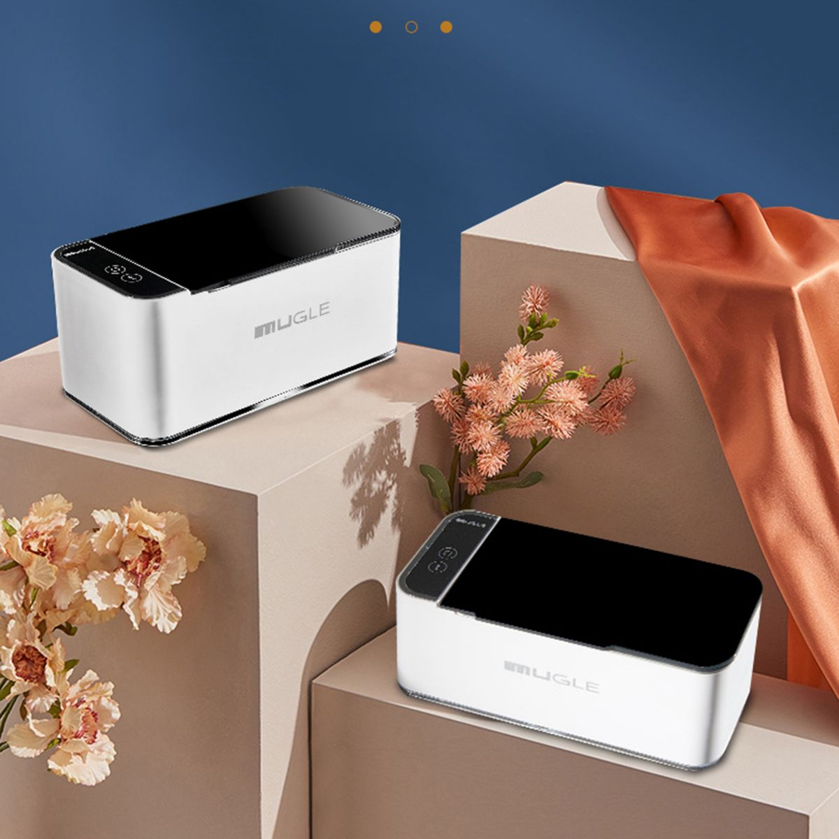 Zwei weiße und schwarze Ultraschallreiniger mit schwarzen Deckeln, auf weißen Blöcken mit Blumen und orangefarbenem Tuch.