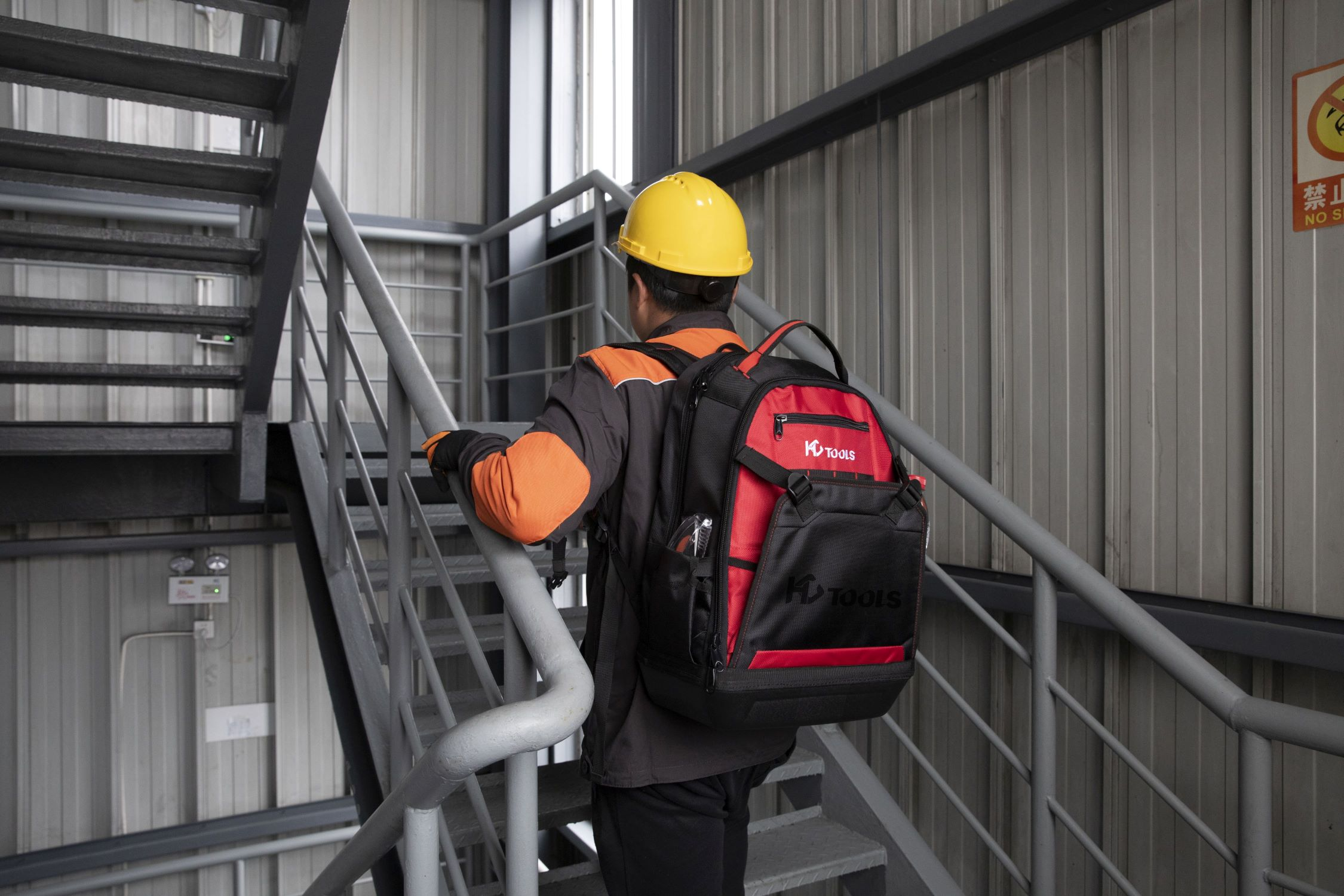 Ein Arbeiter mit gelbem Helm und Rucksack geht eine Metalltreppe in einem Industriegebiet hinauf.