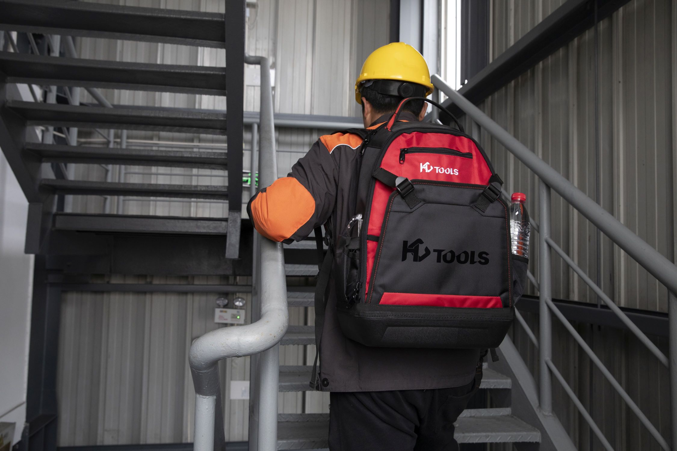 Eine Person in einer orange-schwarzen Jacke und einem gelben Helm geht eine Treppe hinauf und trägt einen Rucksack.