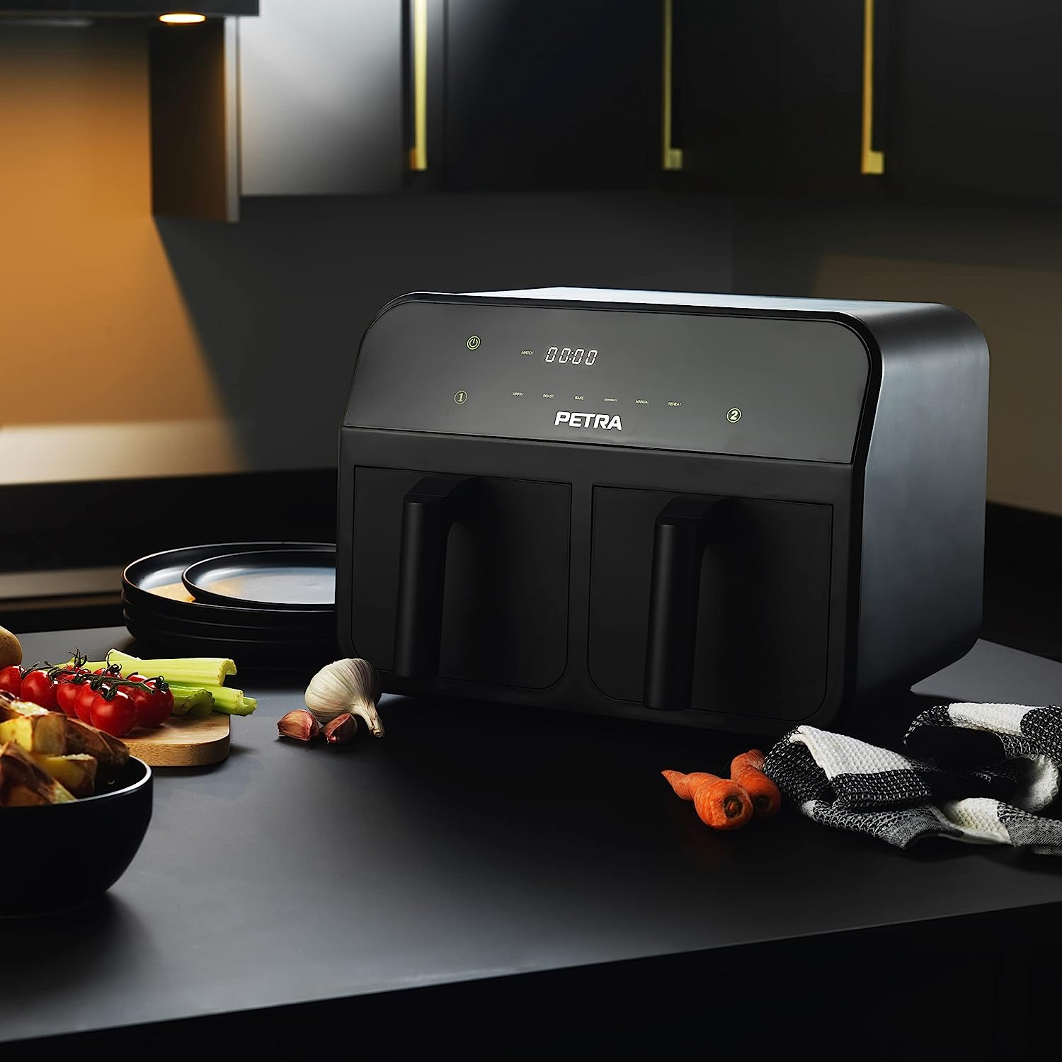 Friggitrice ad aria nera su un piano di lavoro nero con verdure e piatti sullo sfondo.