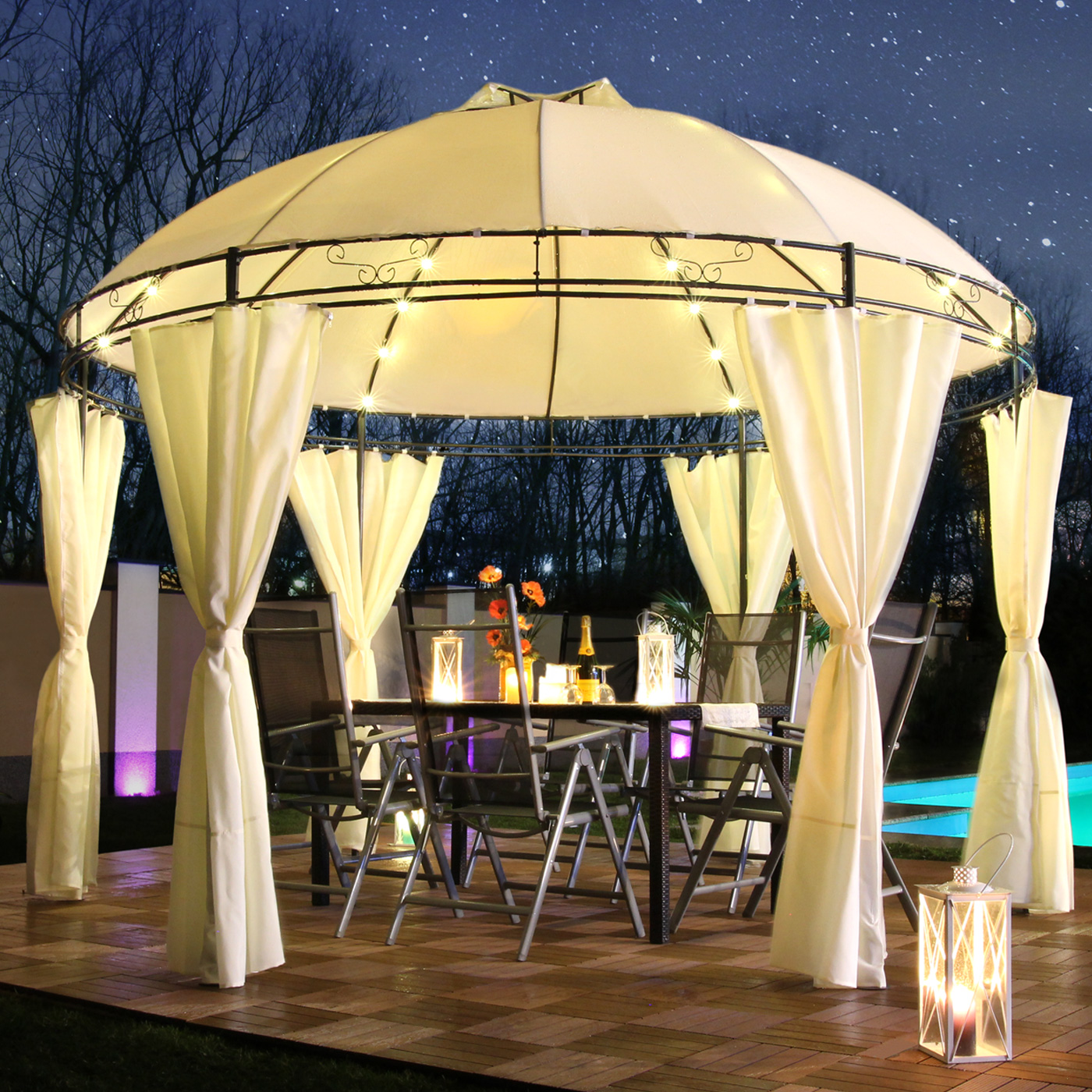 Pavillon bei Nacht mit Lichtern, Tisch, Stühlen und Vorhängen. Bäume im Hintergrund. Lichter sind an.