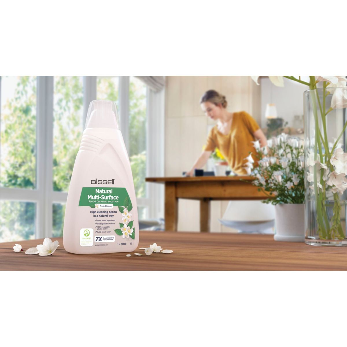Een fles Bissell reinigingsoplossing staat op een houten tafel. Een persoon maakt een tafel schoon op de achtergrond. Er staan ook bloemen in een vaas.