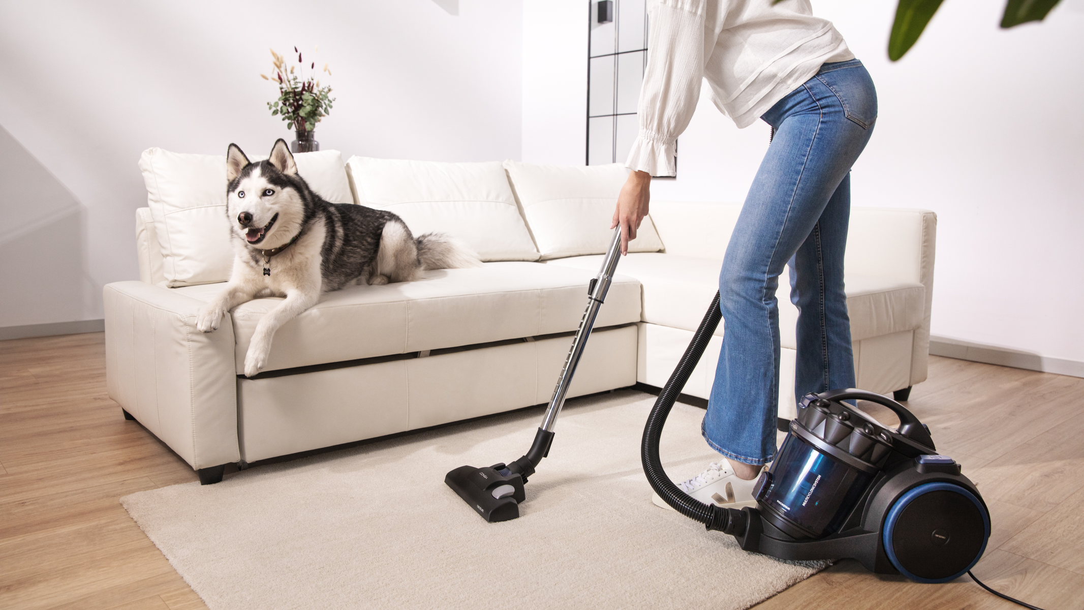 Kobieta odkurza dywan, pies jest na kanapie w tle.