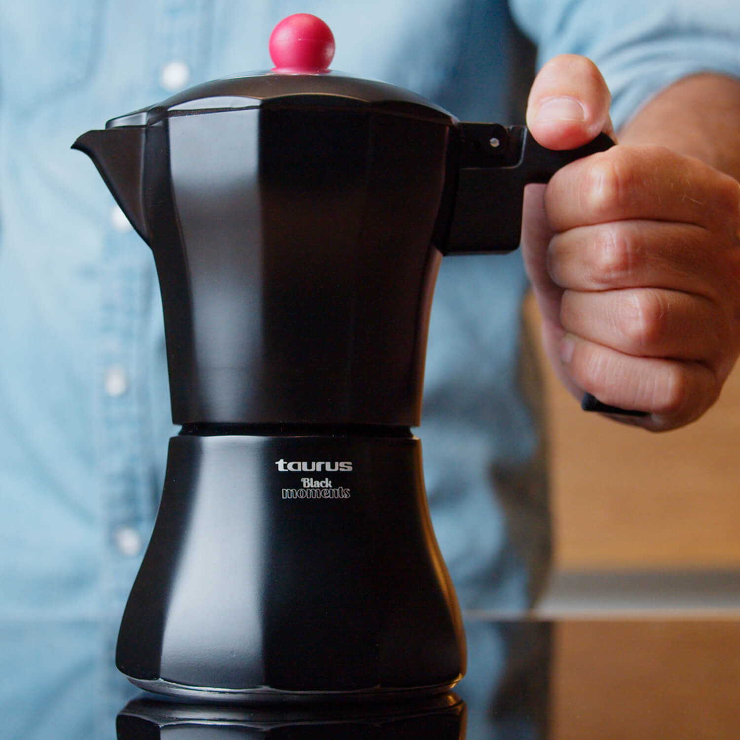 Cafetera exprés negra en una mano. Tiene un botón rojo y la marca Taurus.