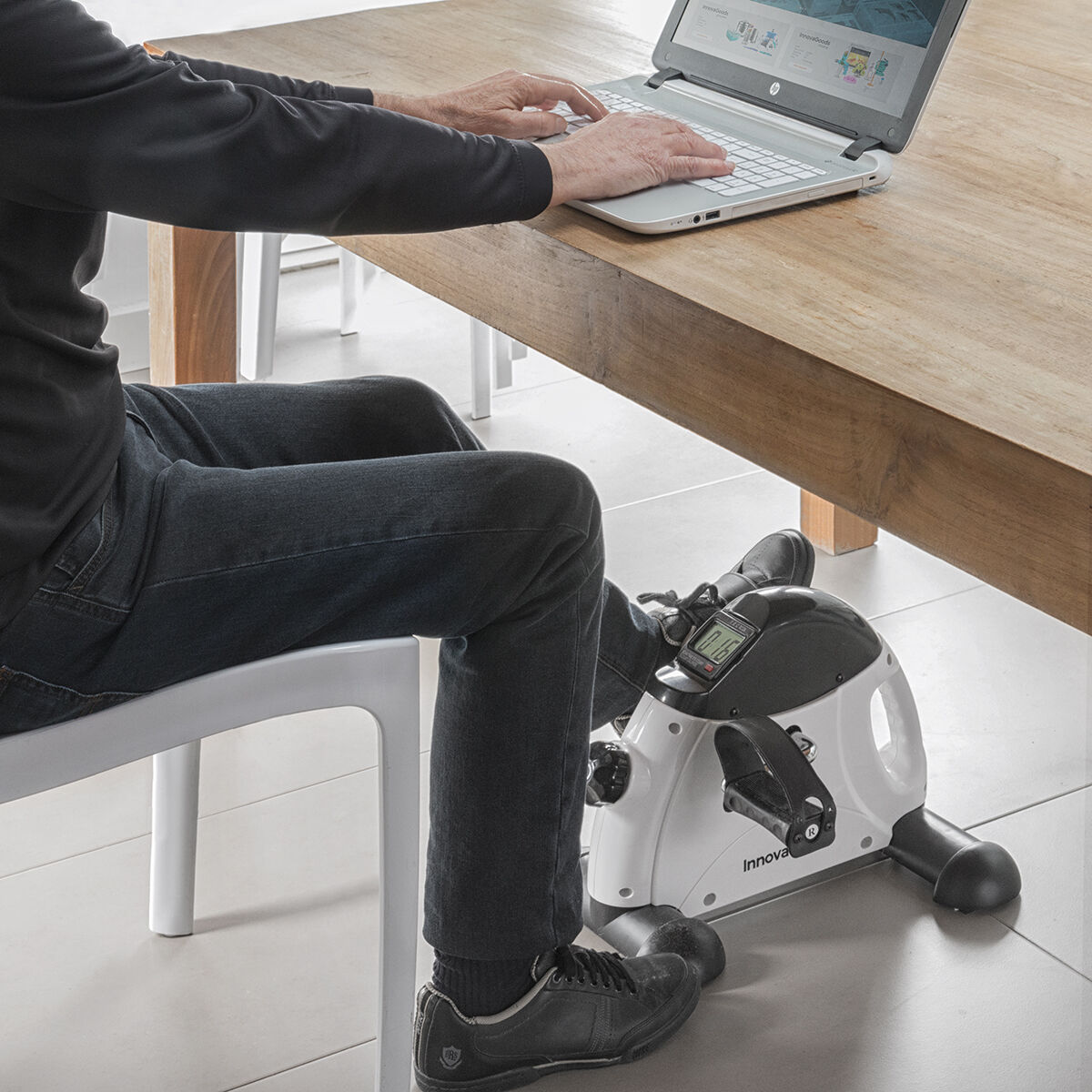 Person nutzt Mini-Heimtrainer unter einem Tisch mit Laptop. Die Szene ist innen.