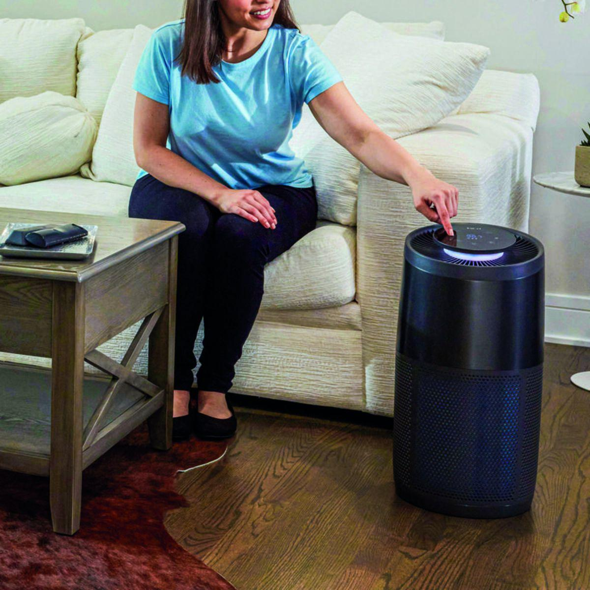 Donna seduta su un divano, aziona un purificatore d'aria. Tavolo con oggetti in primo piano.