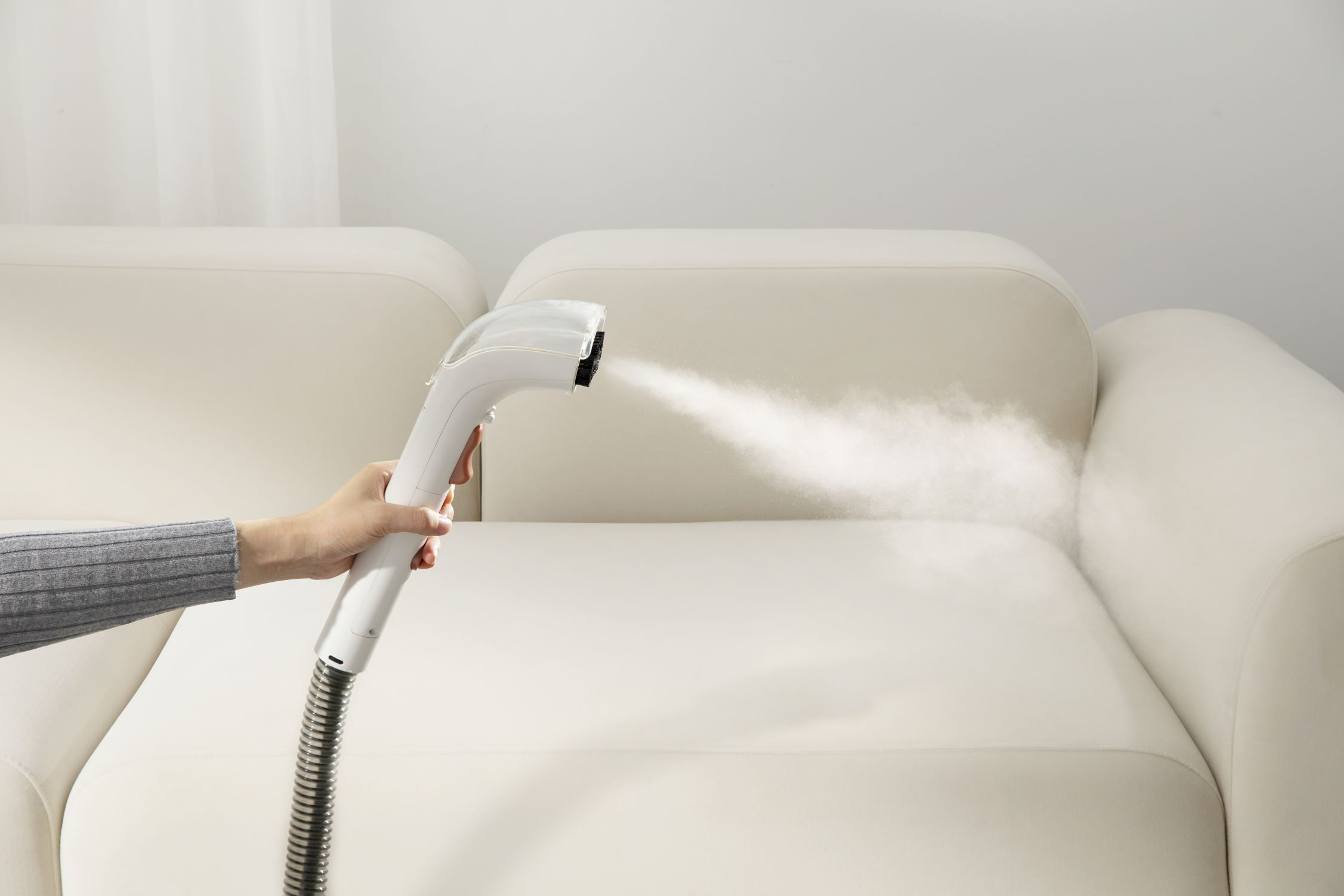 Eine Hand hält einen weißen Dampfreiniger über einem weißen Sofa und gibt Dampf ab.