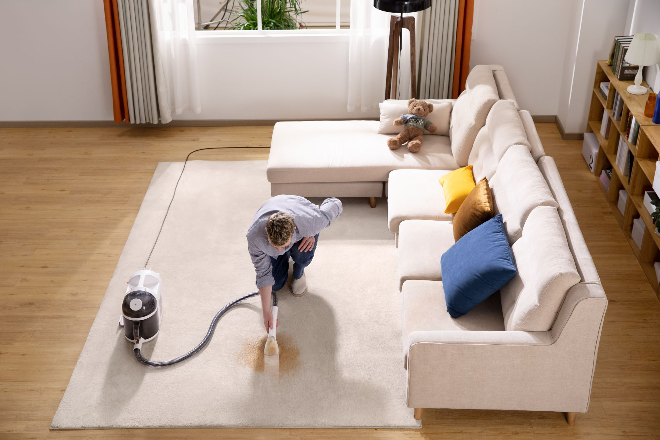 Mann, der einen Teppich saugt. Beiger Teppich, beiges Sofa und dunkler Holzboden. Draufsicht.