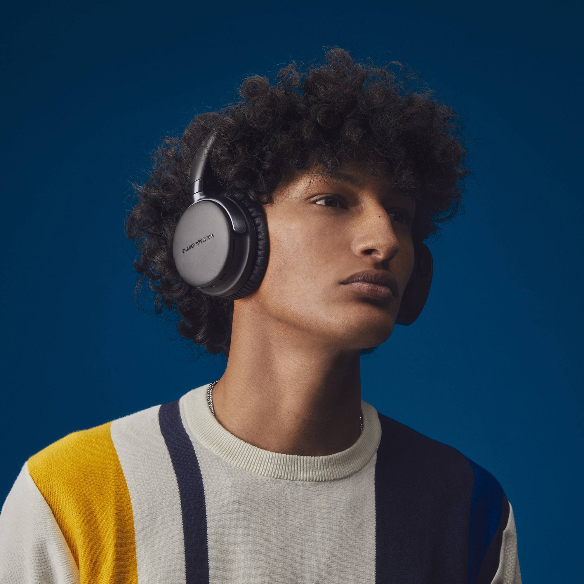 Osoba nosząca czarne słuchawki na niebieskim tle, w pasiastym swetrze. Osoba ma kręcone włosy.