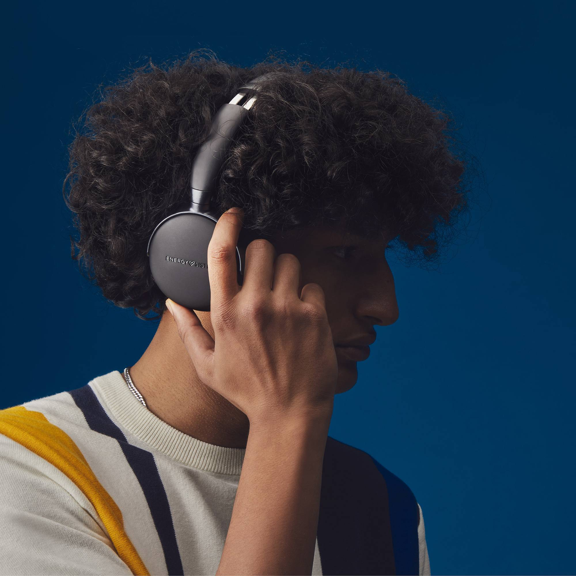 Osoba z kręconymi włosami nosząca czarne słuchawki. Osoba patrzy w kamerę, a tło jest niebieskie.