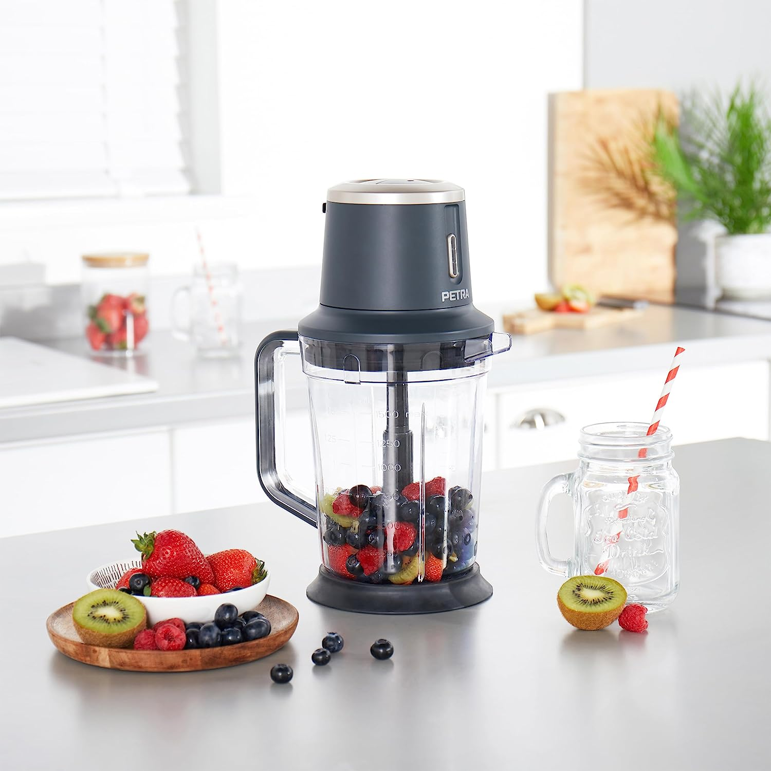 Ein Mixer mit verschiedenen Früchten, ein Glas und Früchte auf einem Holzbrett auf einer Küchentheke.