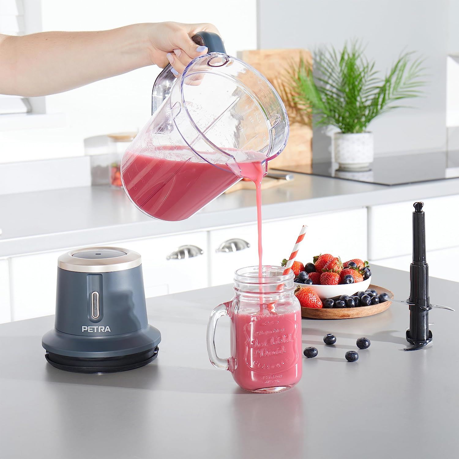 Eine Person gießt pinkfarbenen Smoothie aus einem Mixer in ein Glas mit Strohhalm, Erdbeeren und Blaubeeren.