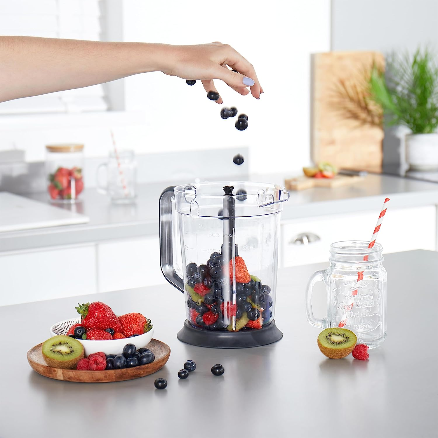 Eine Hand lässt Blaubeeren in einen Mixer fallen. Der Mixer enthält Früchte und steht auf einer Theke.