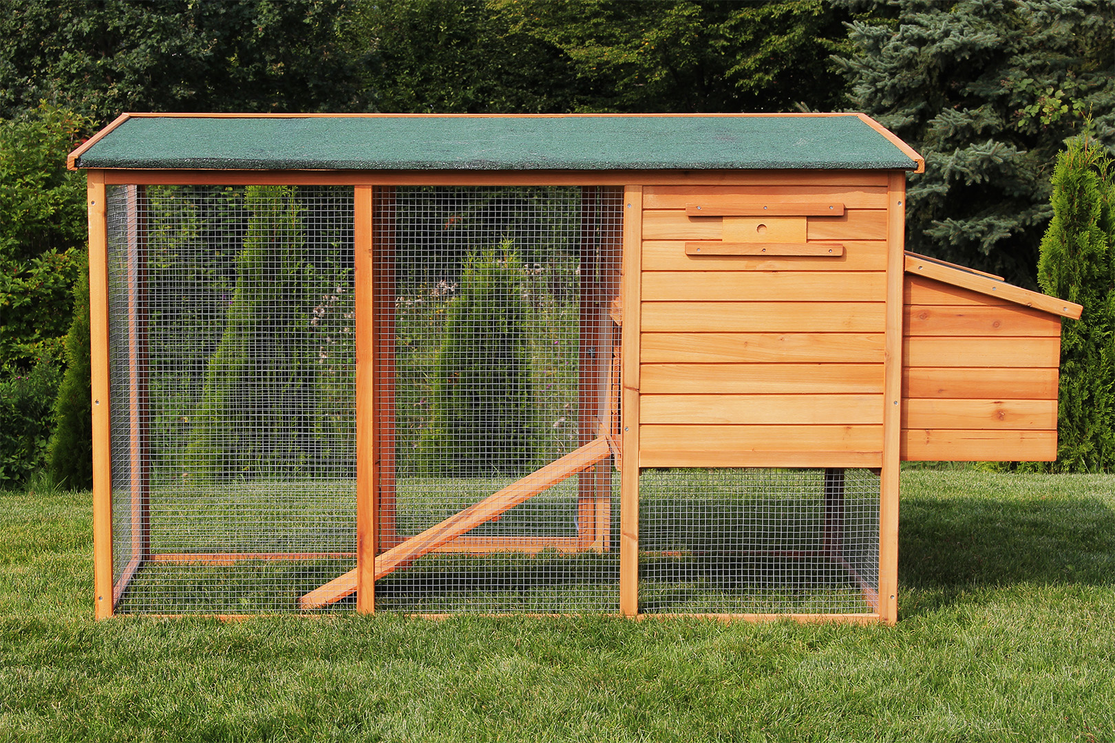 Hühnerstall aus Holz mit Drahtgitterseiten, grünem Dach und Gras im Vordergrund.