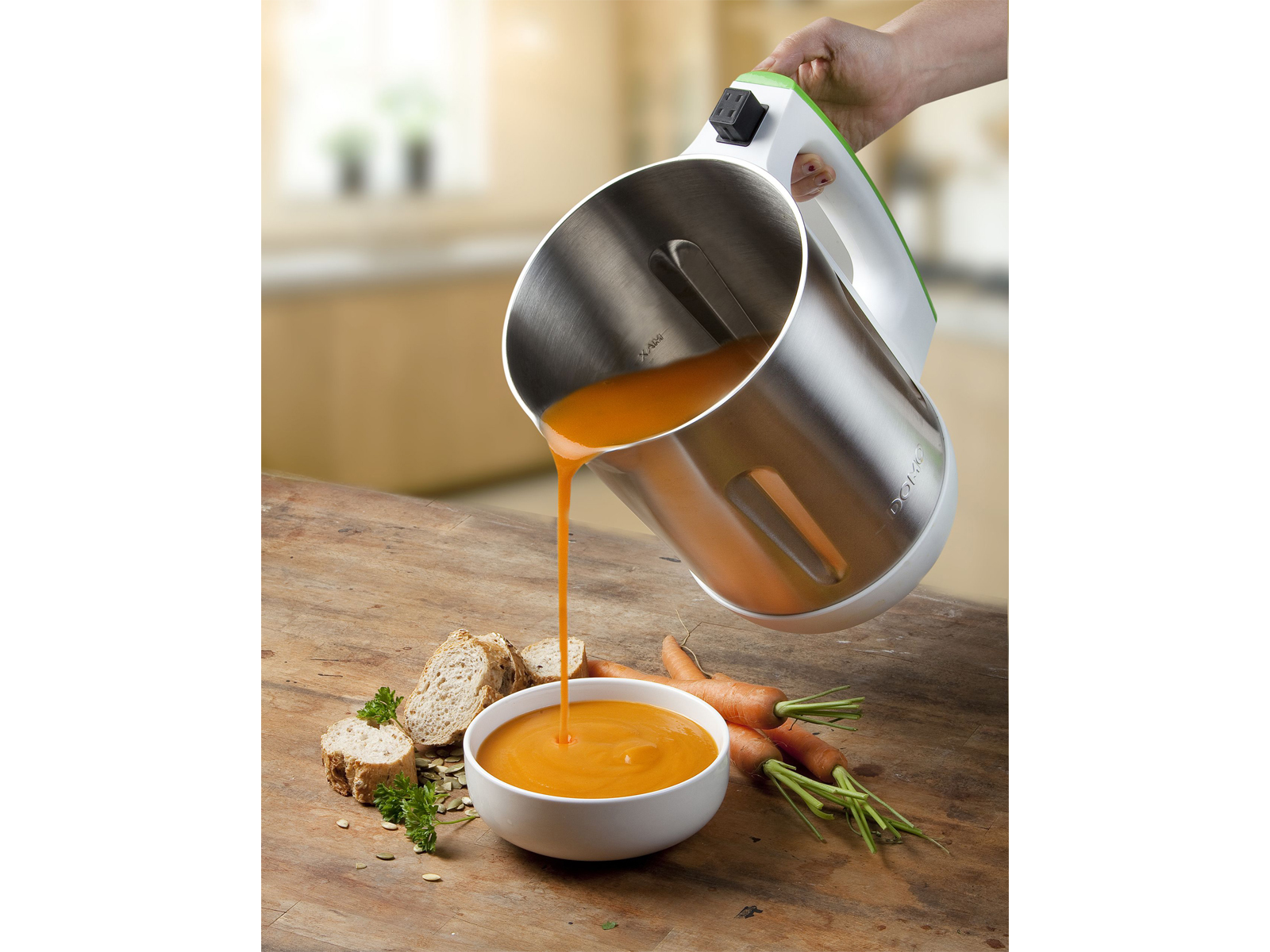Una persona versa zuppa arancione da un produttore di zuppa in una ciotola, accanto a pane e carote.