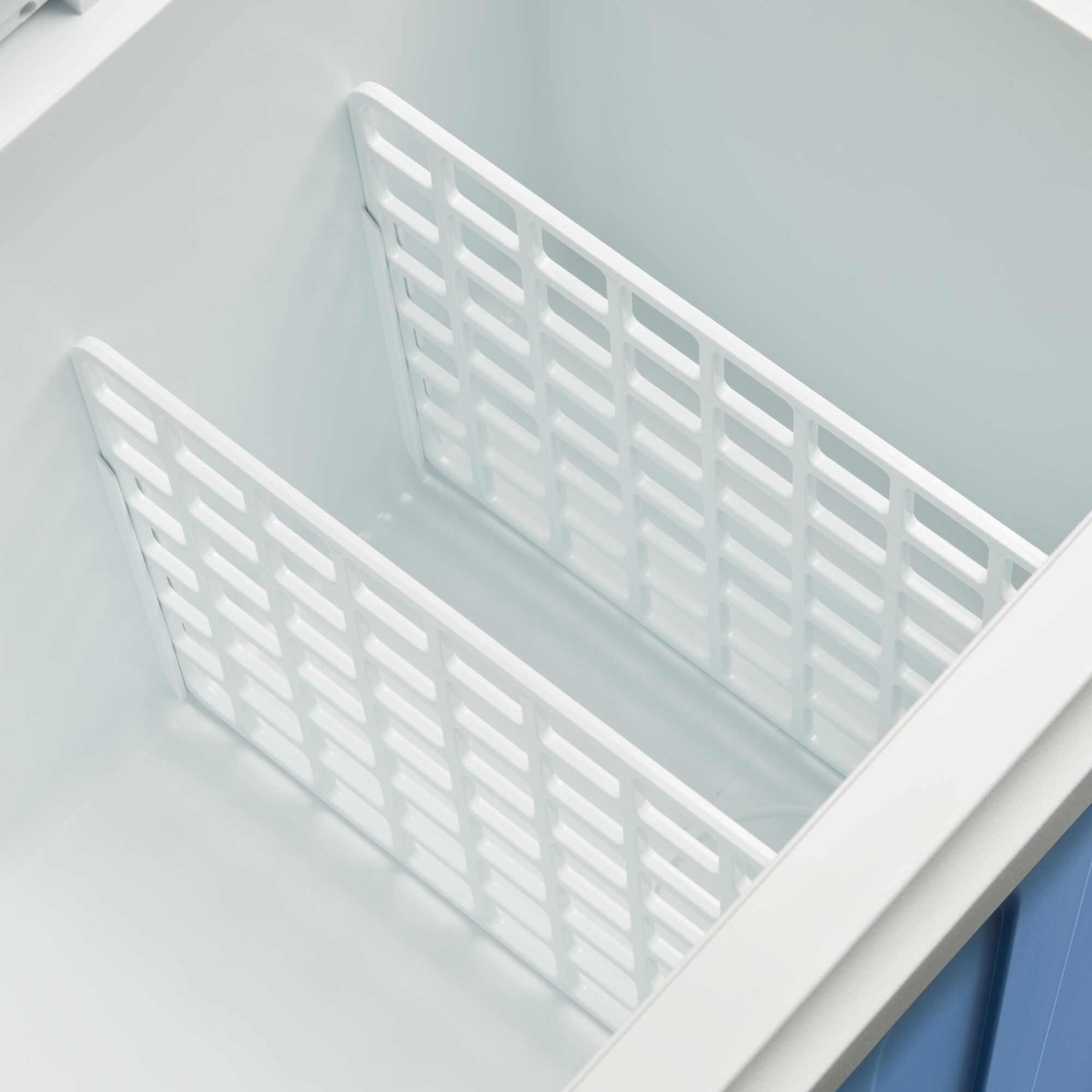 Białe wnętrze zamrażarki z dwoma białymi przegrodami kratowymi. Widoczne niebiesko-białe akcenty.