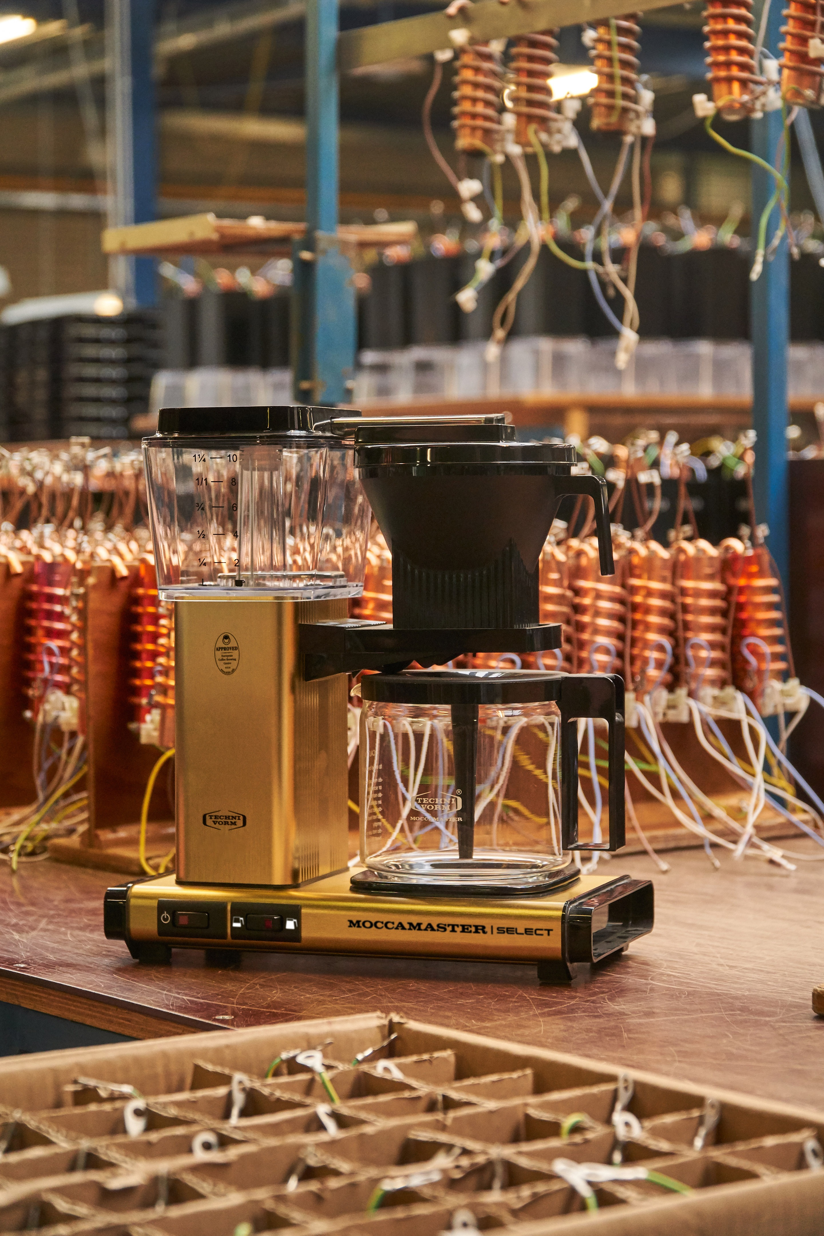 Eine goldene Moccamaster Kaffeemaschine steht auf einem Holztisch in einer Fabrikumgebung mit Kabeln und Spulen.
