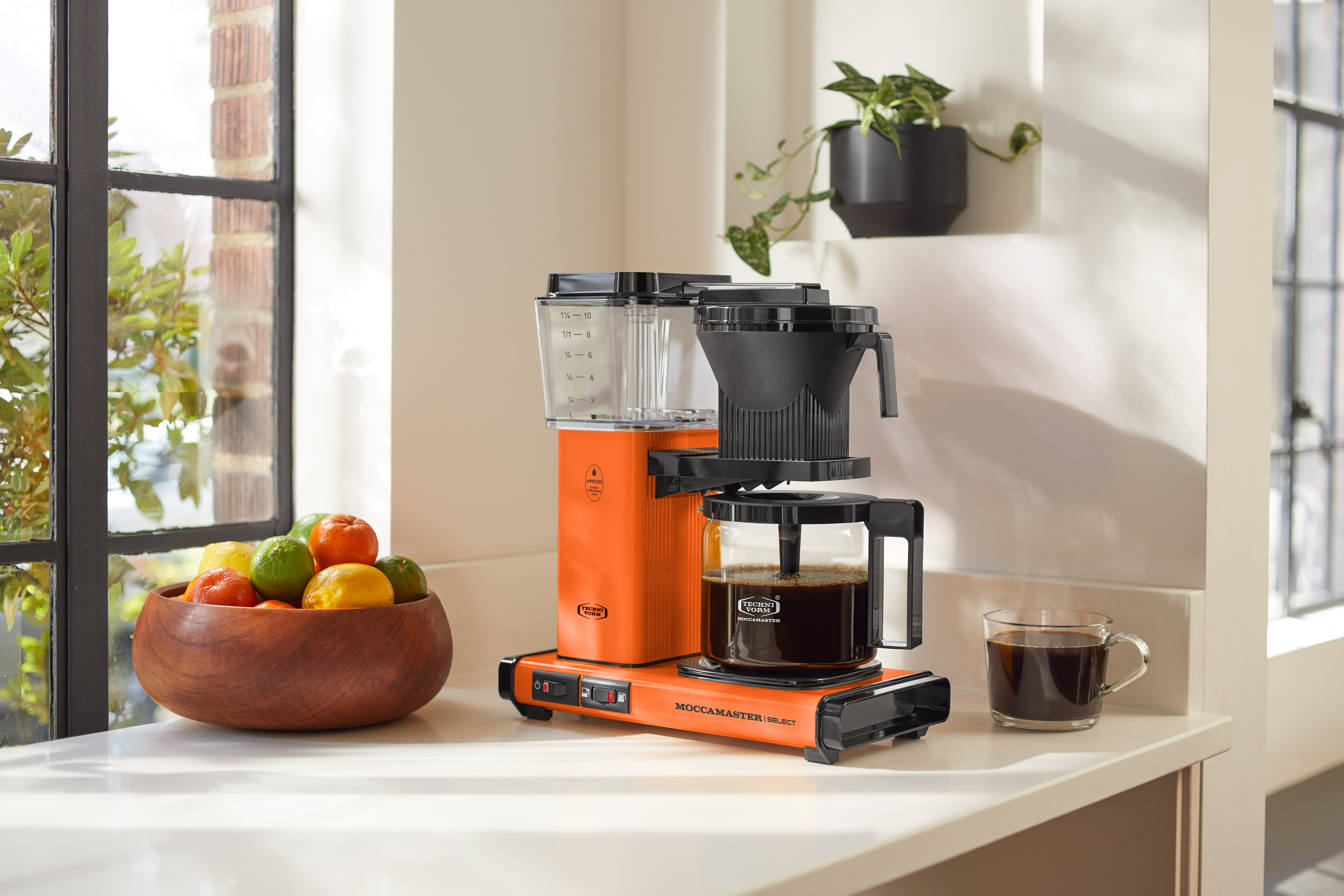 Pomarańczowy ekspres do kawy ze szklaną karafką, filiżanka kawy i miska owoców.