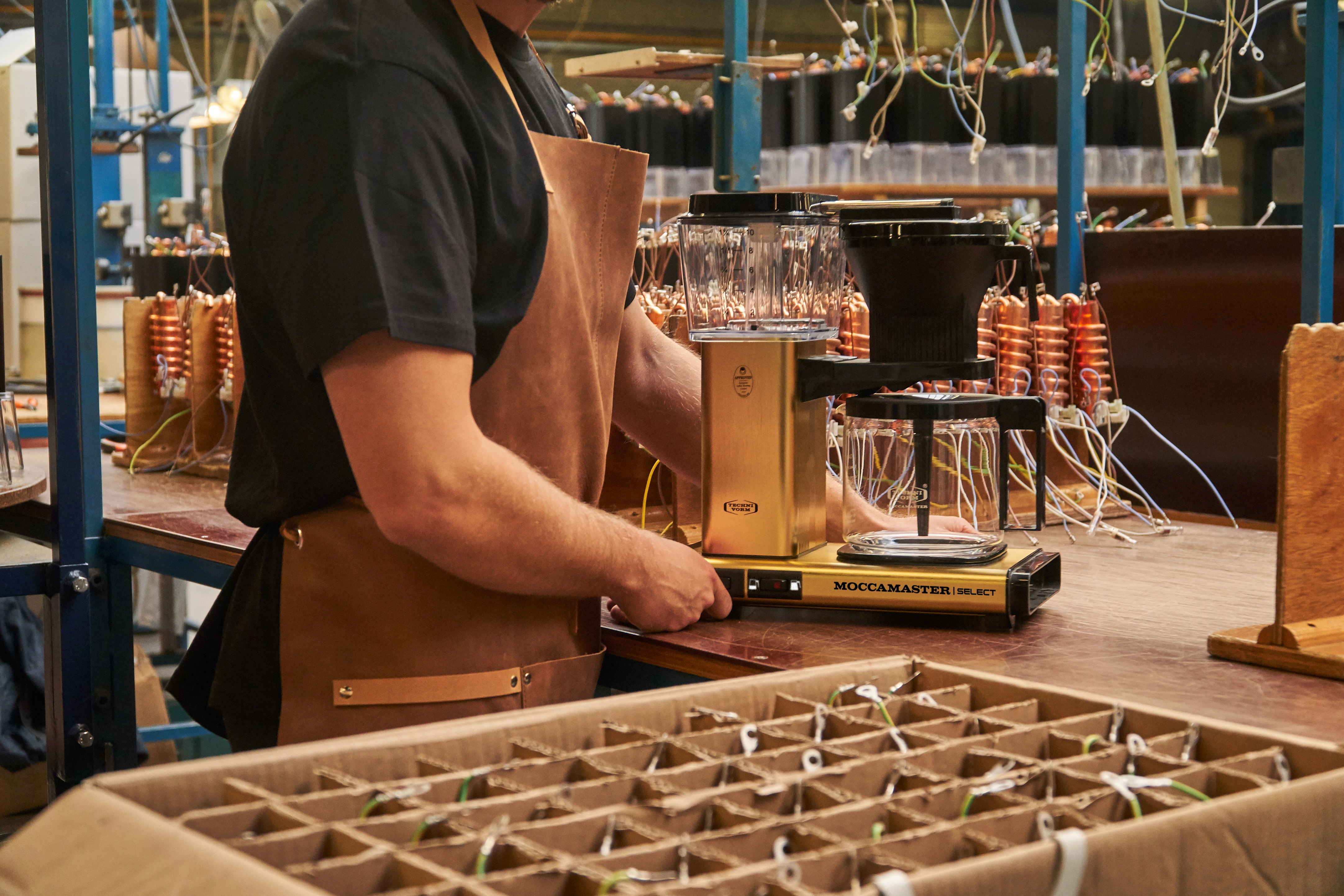 Eine Person, die Kaffee mit einer goldenen Moccamaster Kaffeemaschine zubereitet. Im Hintergrund sind Spulen.