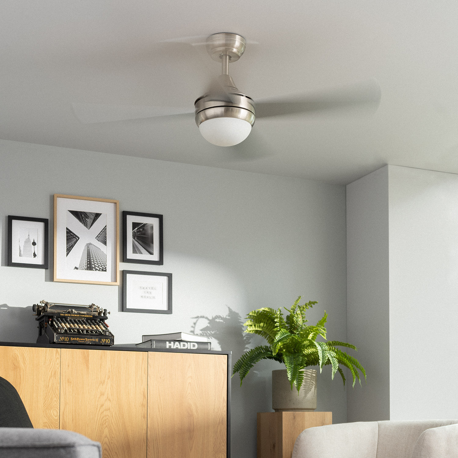 Un ventilatore da soffitto con luce, contro un muro grigio con quadri incorniciati e una felce.