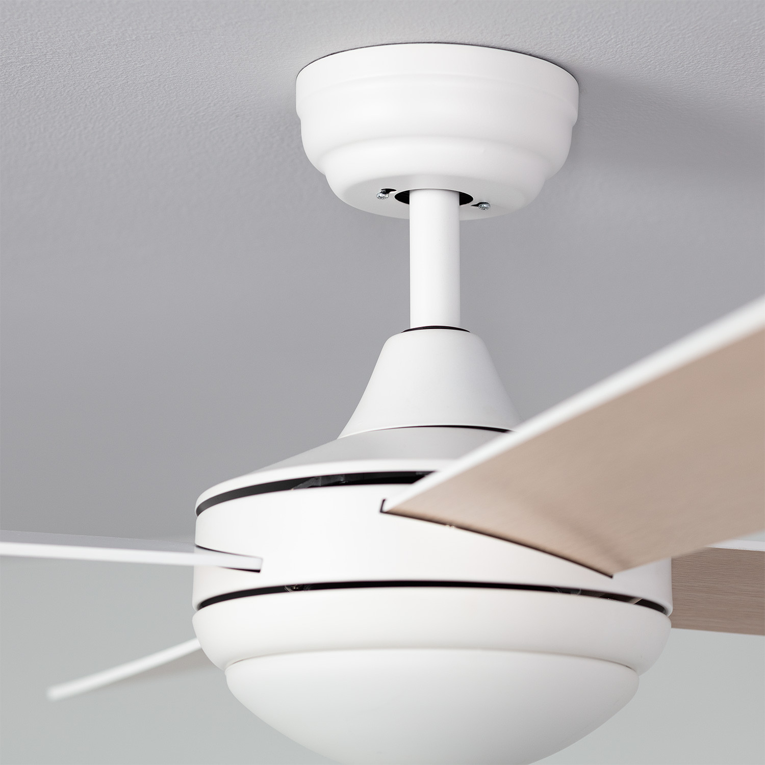 Un ventilatore da soffitto bianco. Le pale sono color legno chiaro. Il ventilatore è contro un muro grigio e un soffitto bianco.