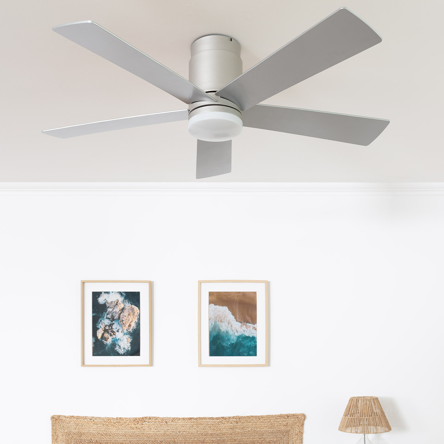 Ventilatore da soffitto con cinque pale argentate. Due quadri incorniciati e una lampada sotto. Soffitto e parete bianchi.