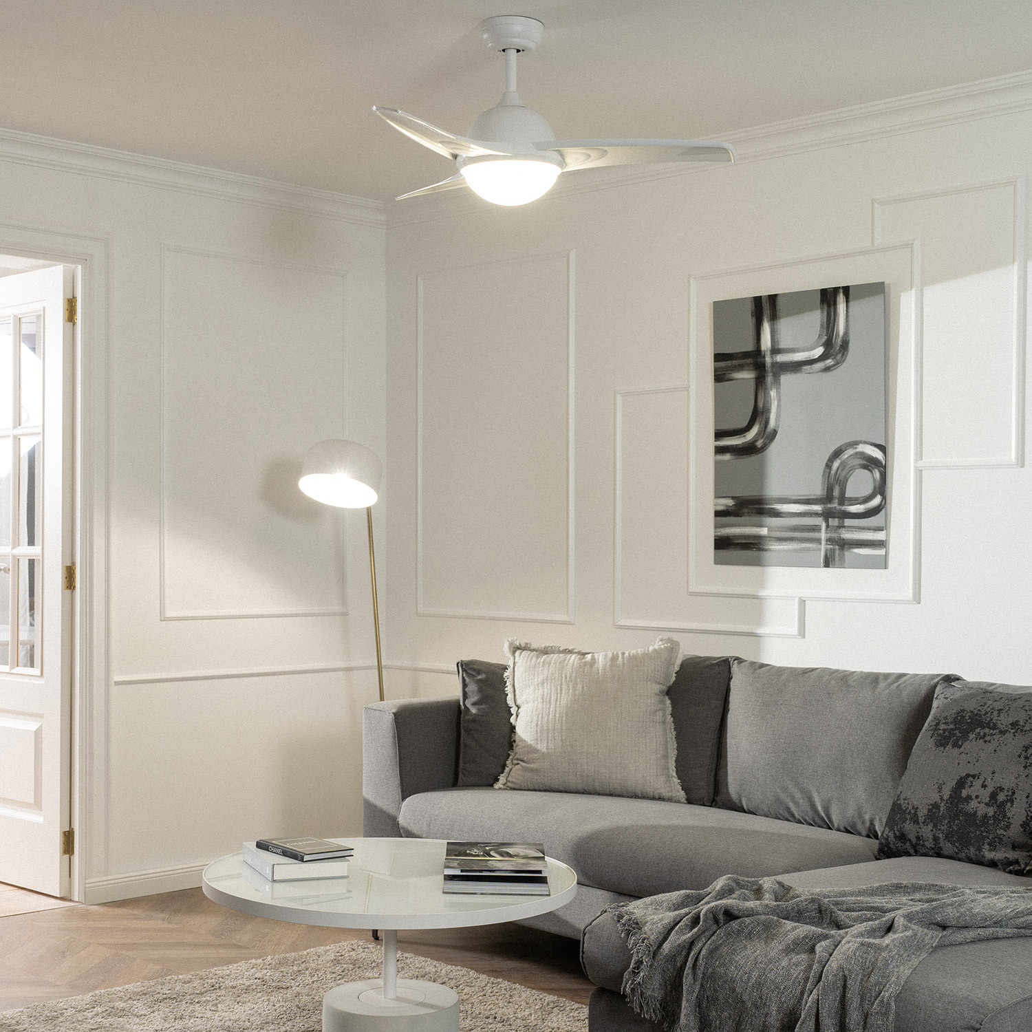 Un soggiorno con ventilatore a soffitto, divano grigio, tavolo bianco e opere d'arte. La luce del sole entra.