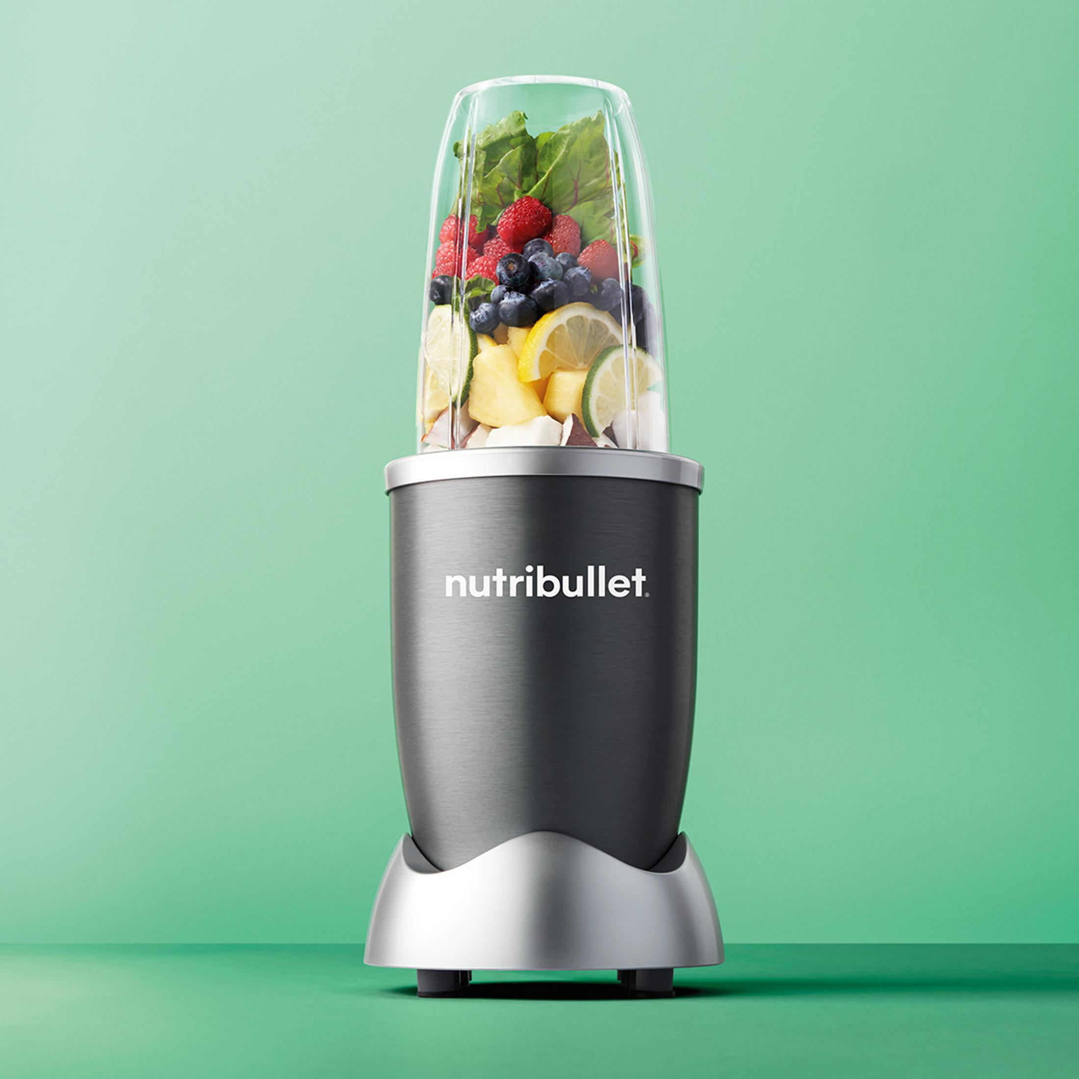 NutriBullet-Mixer mit einem Behälter voller Obst und Gemüse auf grüner Oberfläche.