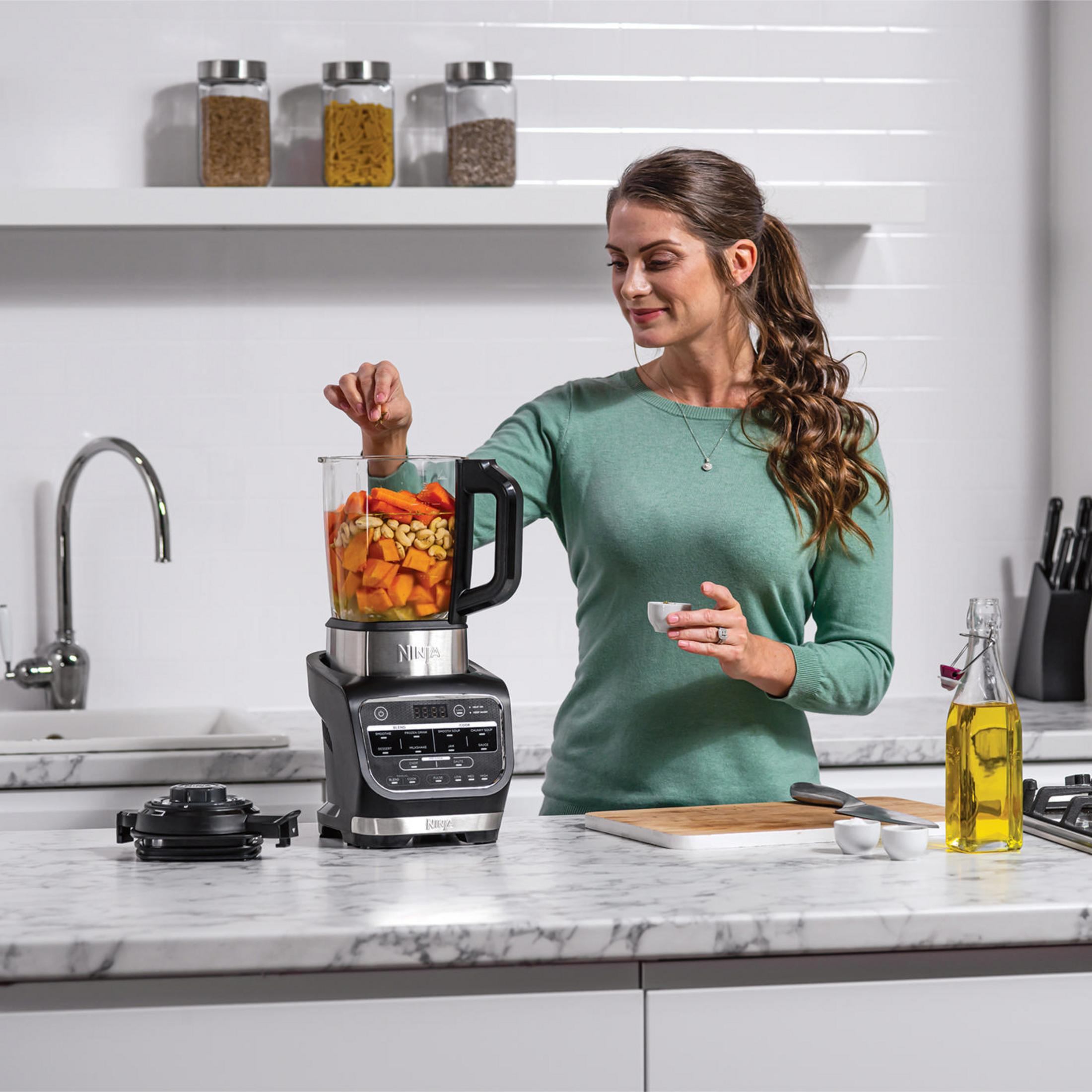 Kobieta dodaje składniki do blendera. Blender stoi na blacie z innymi przyborami kuchennymi.