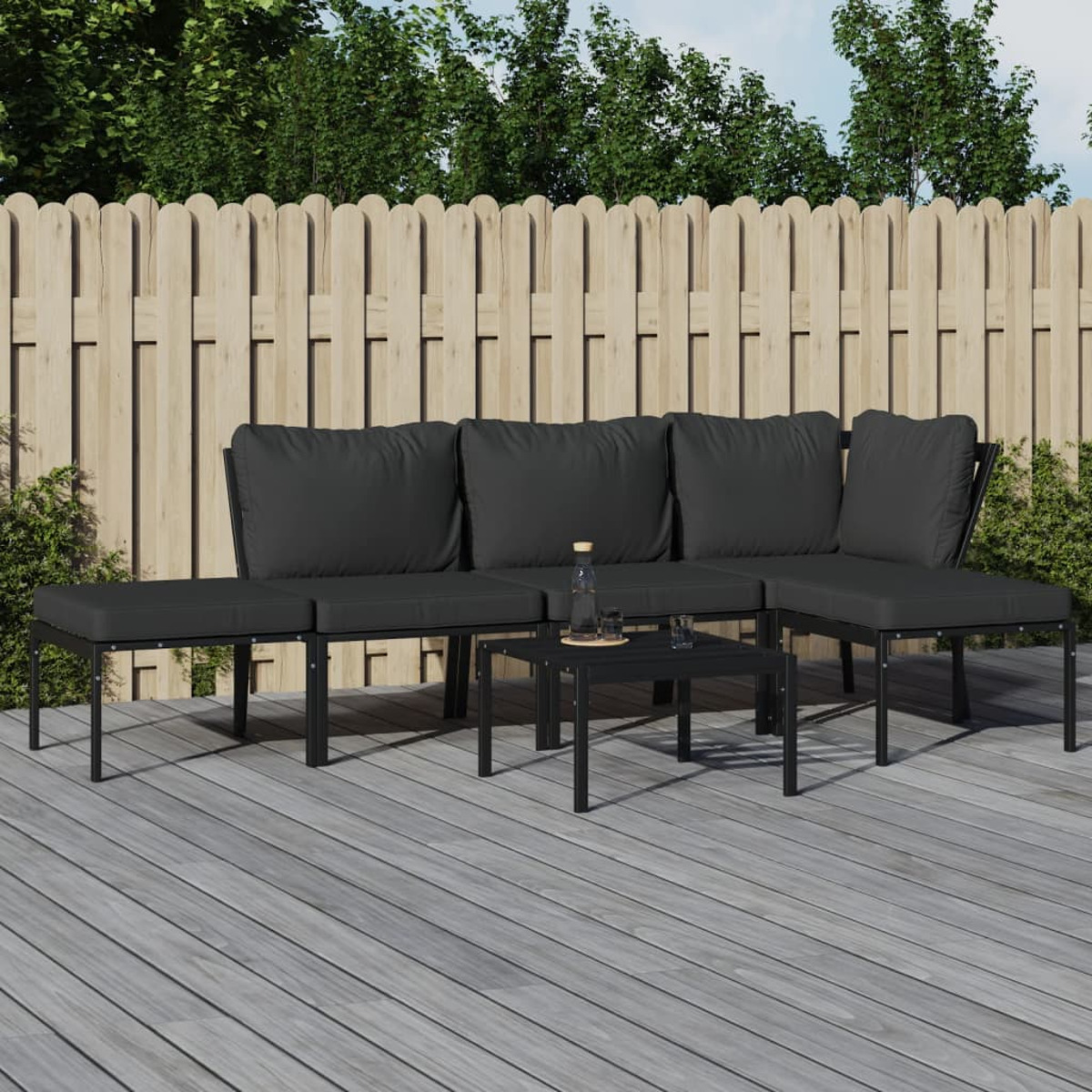 Schwarzes Outdoor-Ecksofa mit dunklen Kissen und kleinem Tisch. Auf Holzdeck, Holzzaun im Hintergrund.